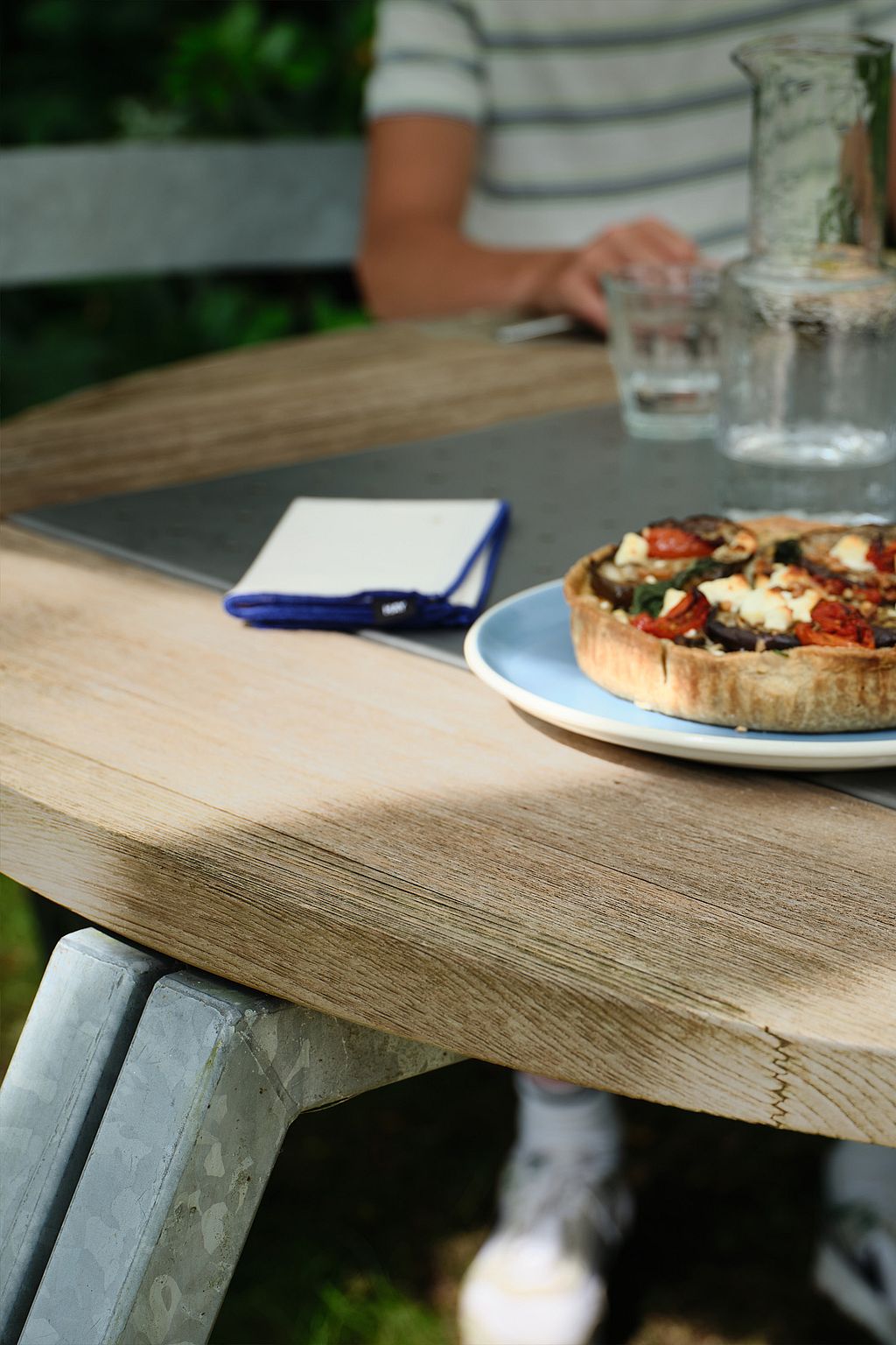 Close-up of a wooden round tabletop with a savory tart on a plate, glasses, and a folded napkin, with a seated person softly blurred behind.