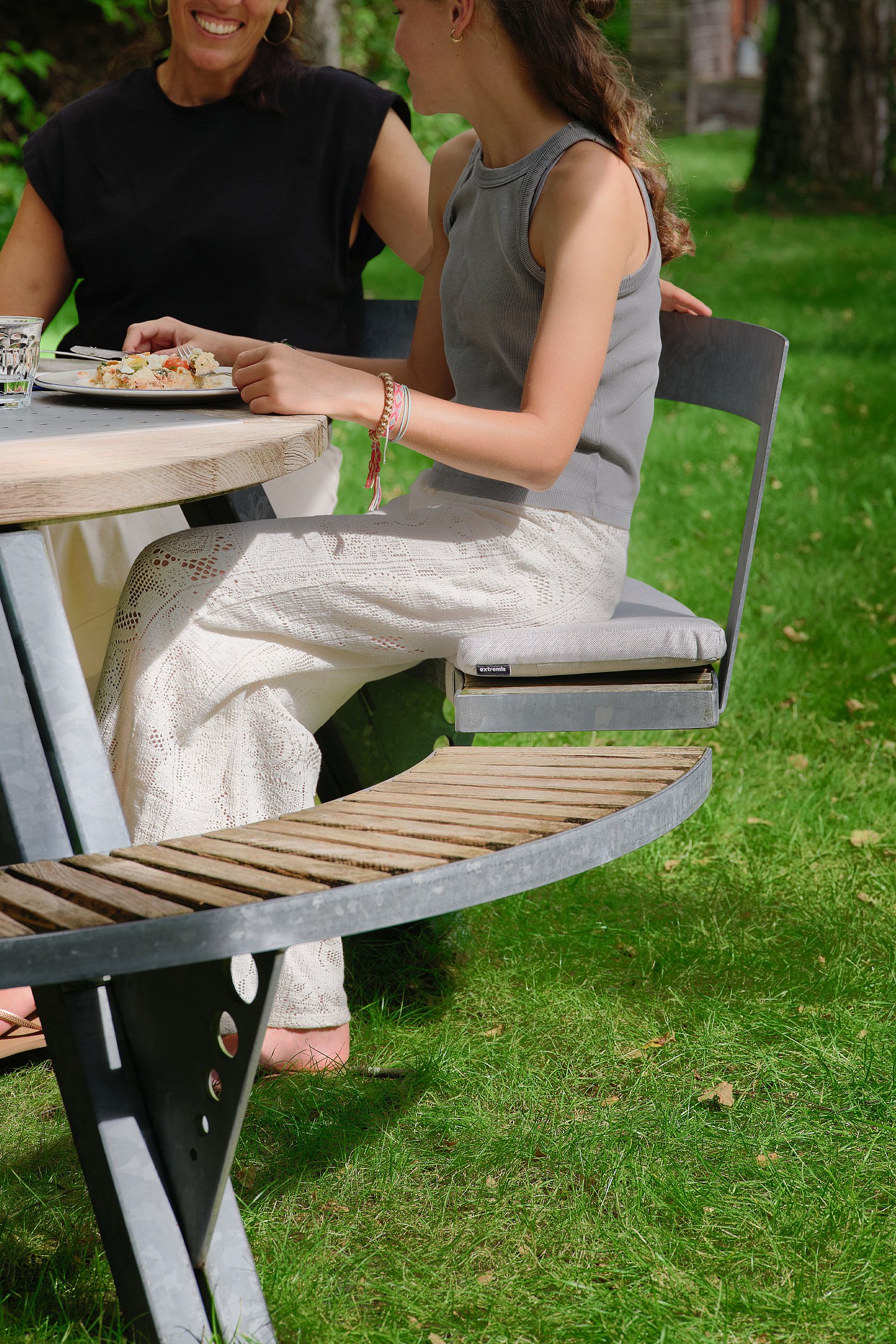 Close-up of two people seated at the Gargantua table, showing the curved wooden bench, raised seat cushion, and galvanized frame.