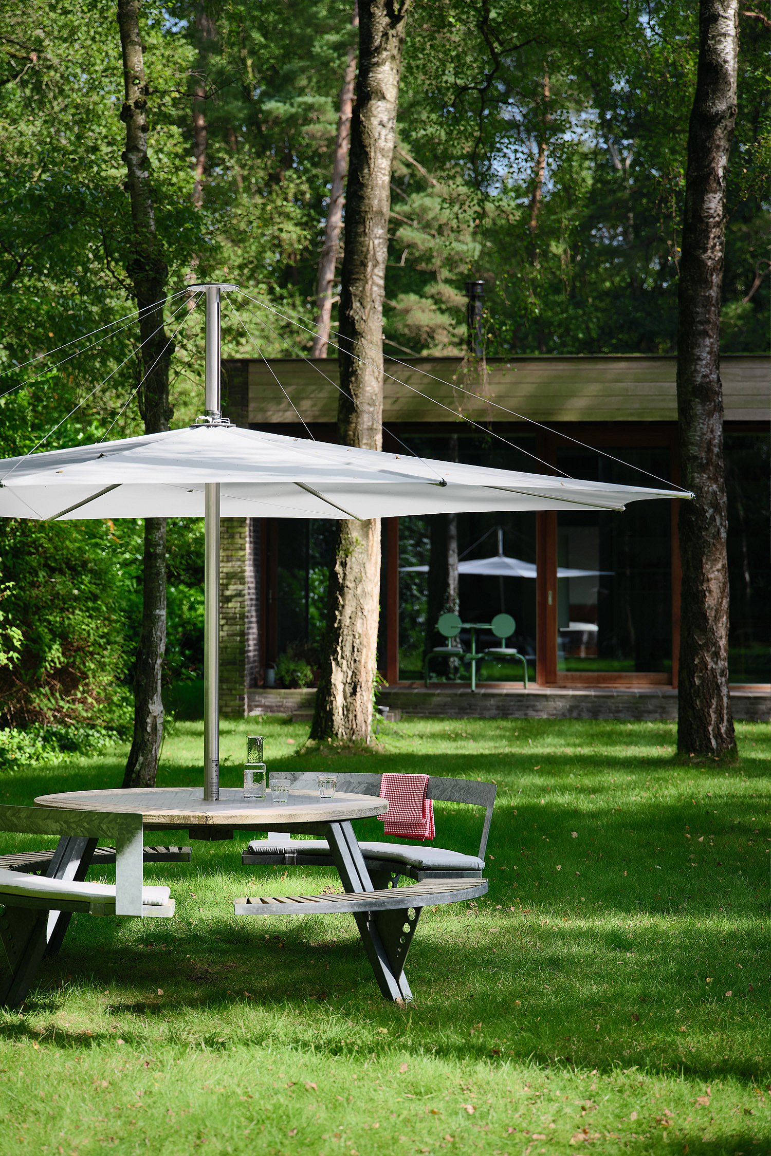 Large white inumbra parasol on a green lawn in front of a low modern house with dark brick walls, broad glass windows, and pale green Bistroo chairs visible inside.