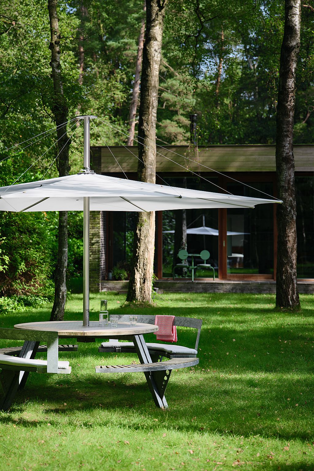 Large white inumbra parasol on a green lawn in front of a low modern house with dark brick walls, broad glass windows, and pale green Bistroo chairs visible inside.