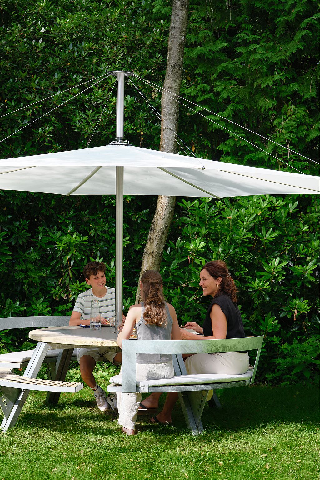 Three people seated around a round outdoor gargantua under a large white inumbra parasol, framed by dense green shrubs and trees.