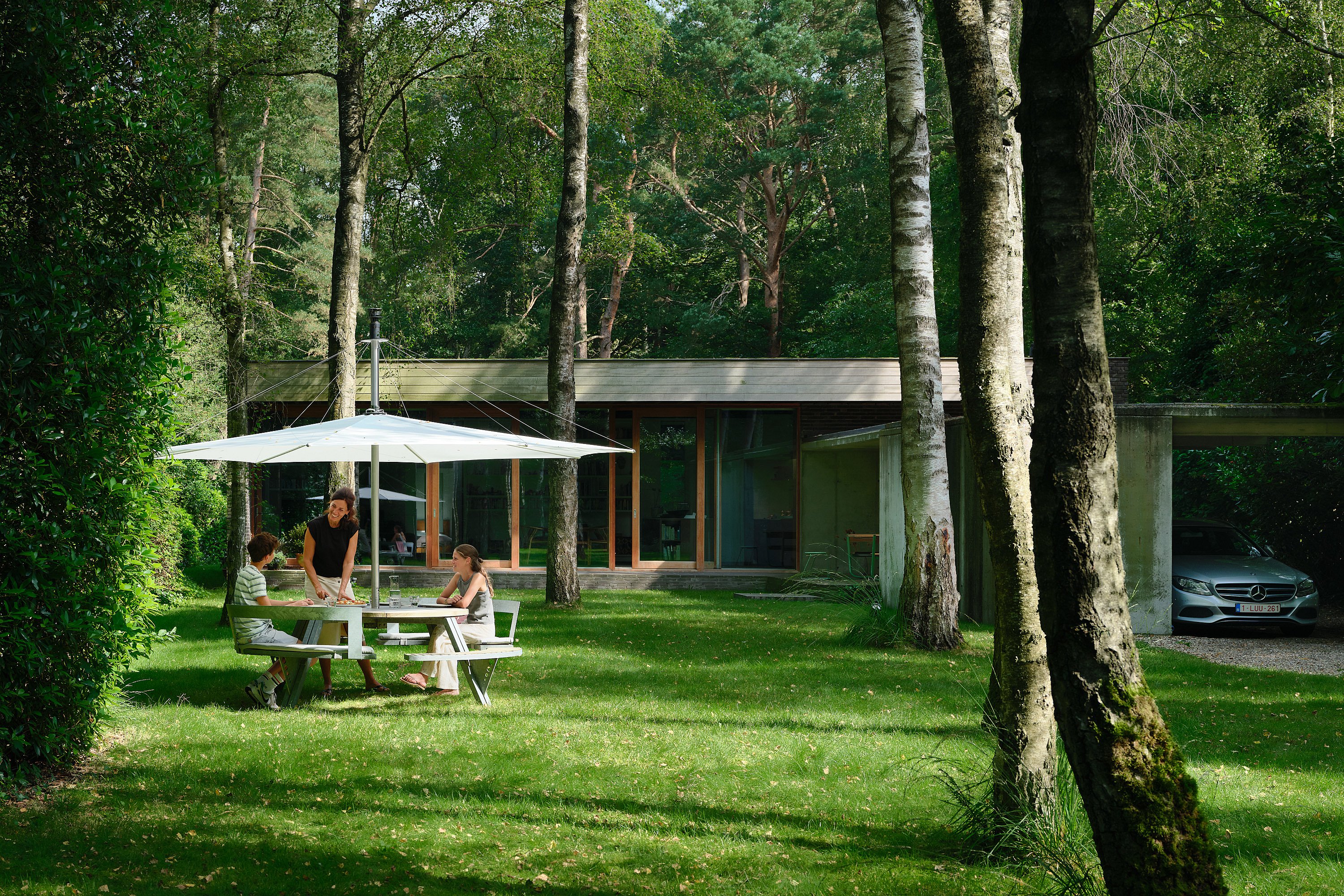 Three people seated around a round outdoor gargantua under a large white inumbra parasol, framed by dense green shrubs and trees.