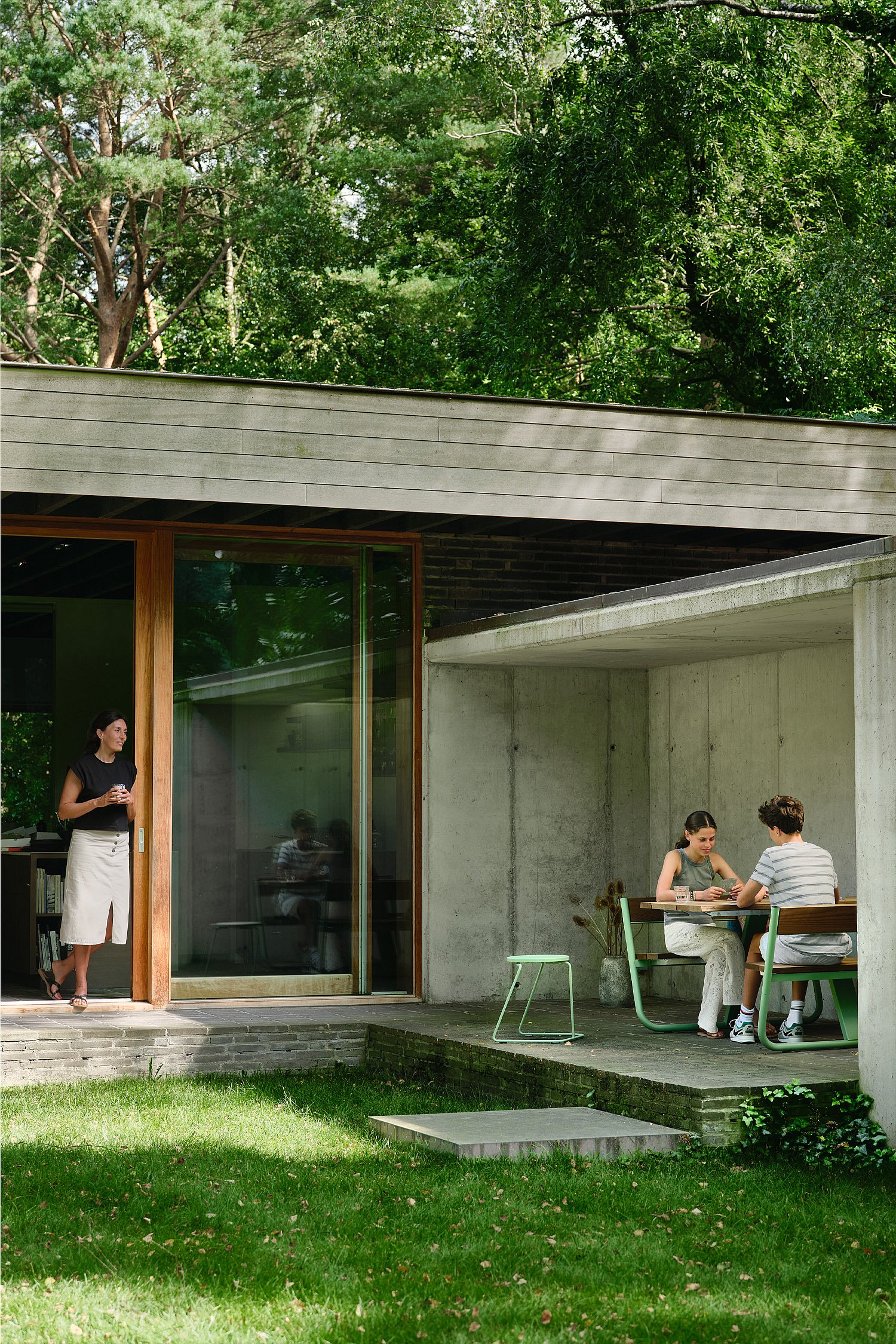 Modern brick-and-concrete house opening onto the garden, with a woman standing by a tall glass door while two people sit together beneath the concrete overhang.
