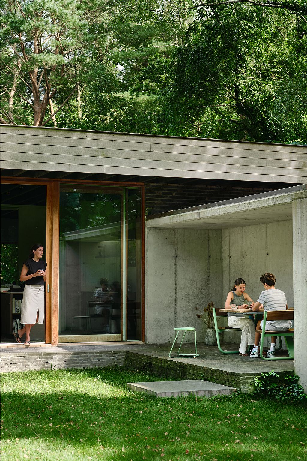 Modern brick-and-concrete house opening onto the garden, with a woman standing by a tall glass door while two people sit together beneath the concrete overhang.