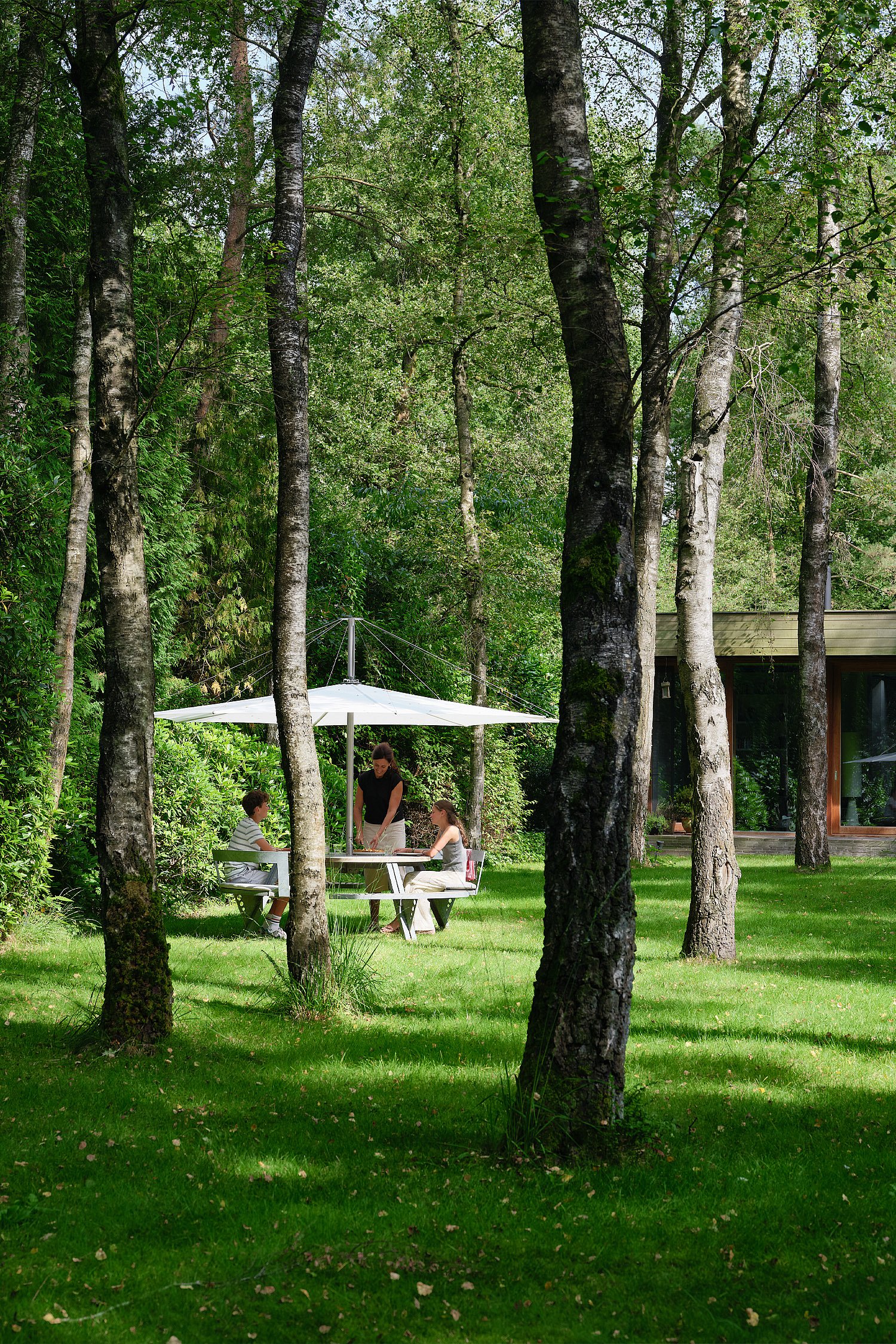 Runder Gargantua-Picknicktisch unter einem großen weißen Sonnenschirm auf einer von hohen Bäumen umgebenen Wiese, mit drei zusammensitzenden Personen neben einem niedrigen modernen Haus im Hintergrund.