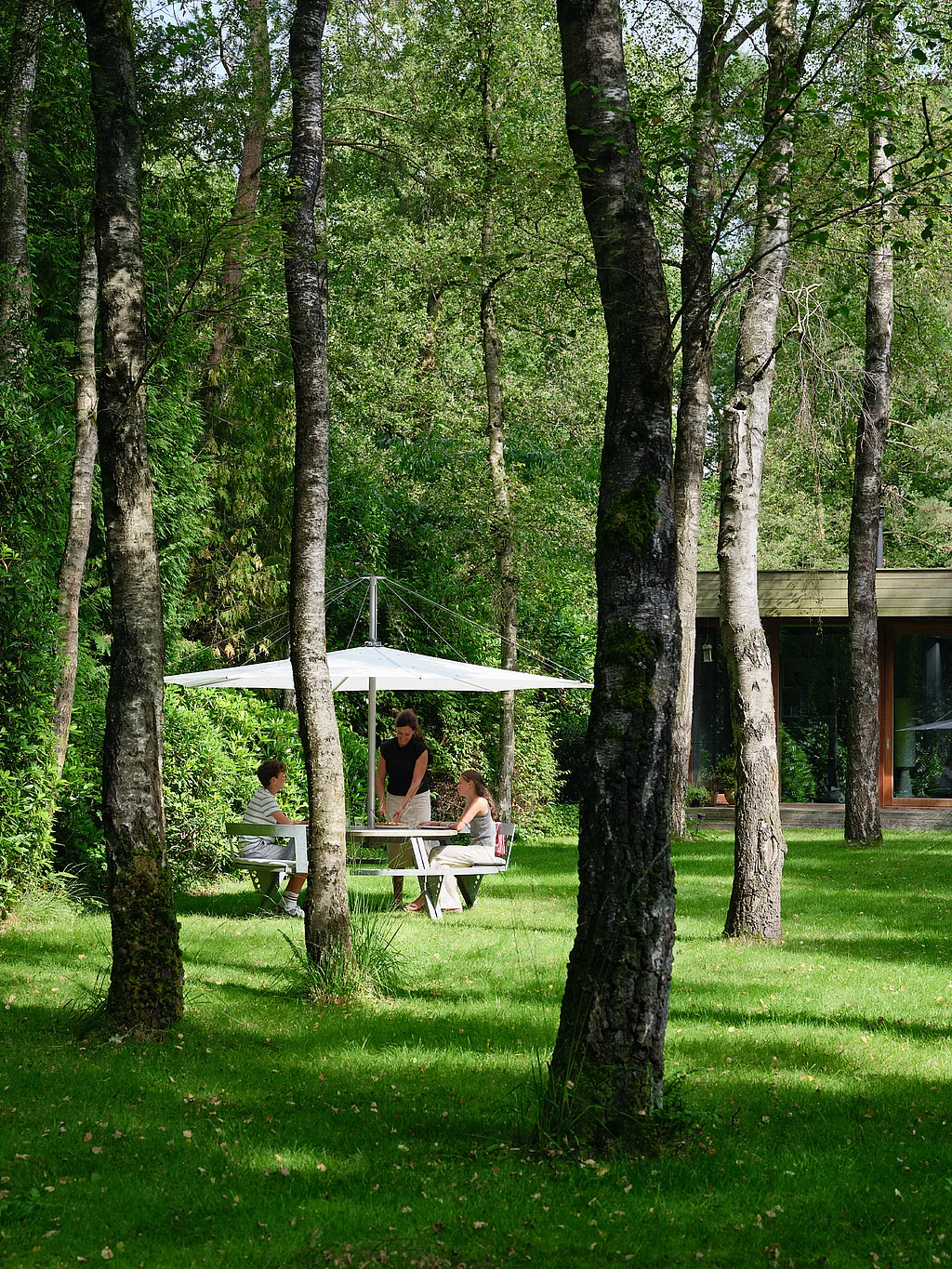 Runder Gargantua-Picknicktisch unter einem großen weißen Sonnenschirm auf einer von hohen Bäumen umgebenen Wiese, mit drei zusammensitzenden Personen neben einem niedrigen modernen Haus im Hintergrund.