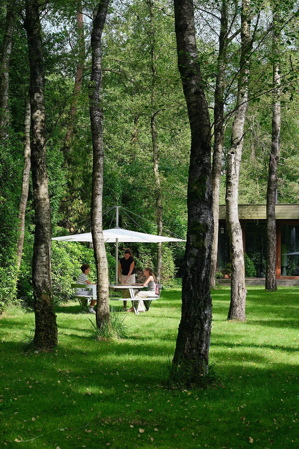 Round Gargantua picnic table under a large white parasol on a lawn surrounded by tall trees, with three people seated together beside a low modern house in the background.