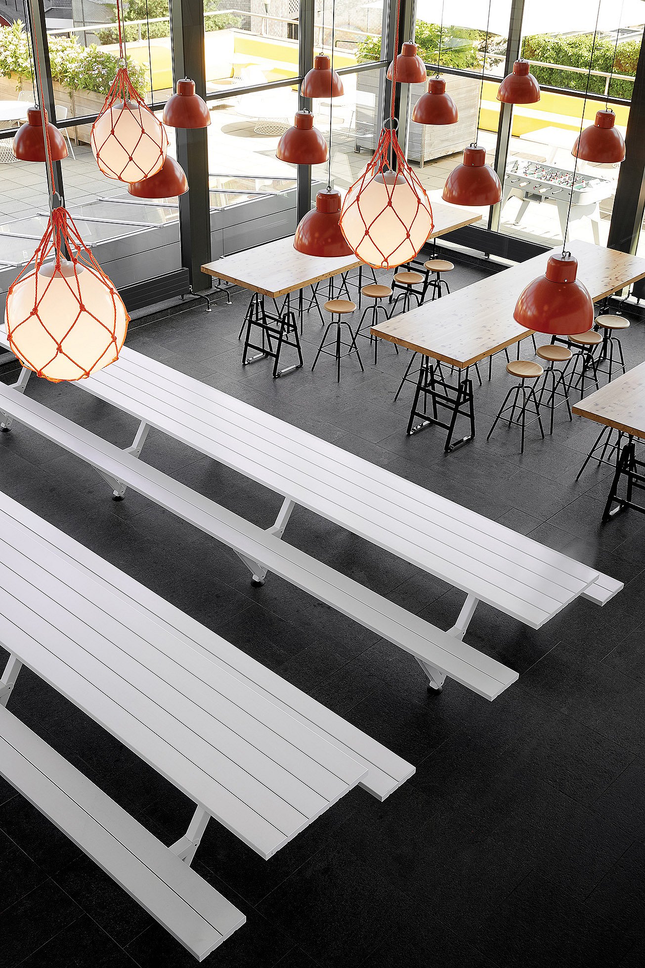 Long white Marina picnic tables in a double-height cafeteria, with large orange pendant lights overhead and floor-to-ceiling windows looking out onto an outdoor terrace.