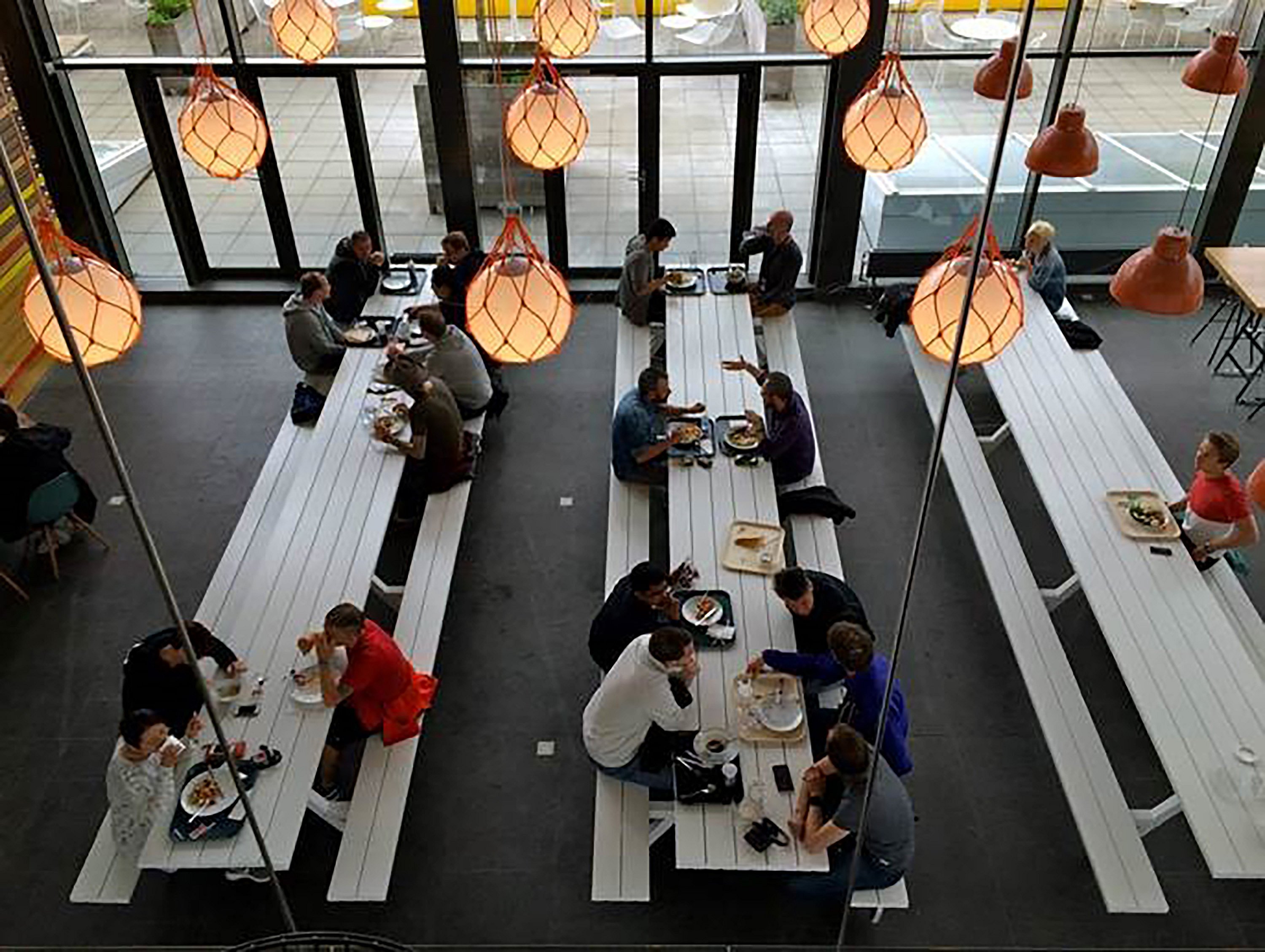 Vue aérienne des employés mangeant et parlant à de longues tables blanches de pique-nique de la Marina sous des lumières suspendues orange brillantes à côté d’un mur de verre pleine hauteur.