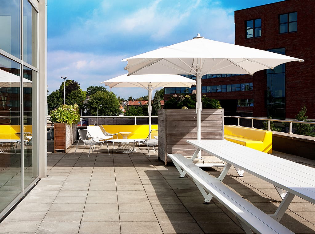 Table de pique-nique White Marina sur une terrasse sur le toit avec de grands parasols blancs, des banquettes intégrées jaunes, des chaises longues, des jardinières et des immeubles de bureaux en arrière-plan.