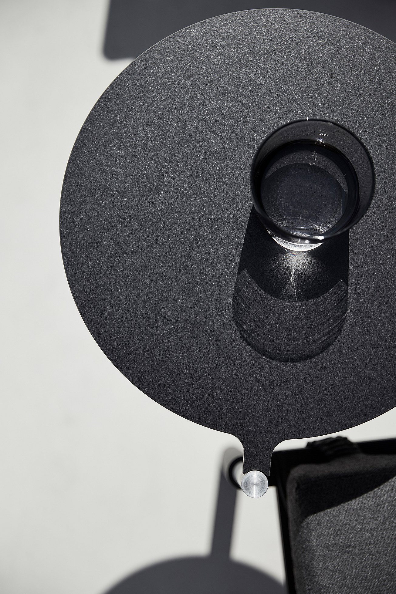 Round dark side table attached to a lounge chair, viewed from above with a clear drinking glass casting a sharp shadow on the tabletop.