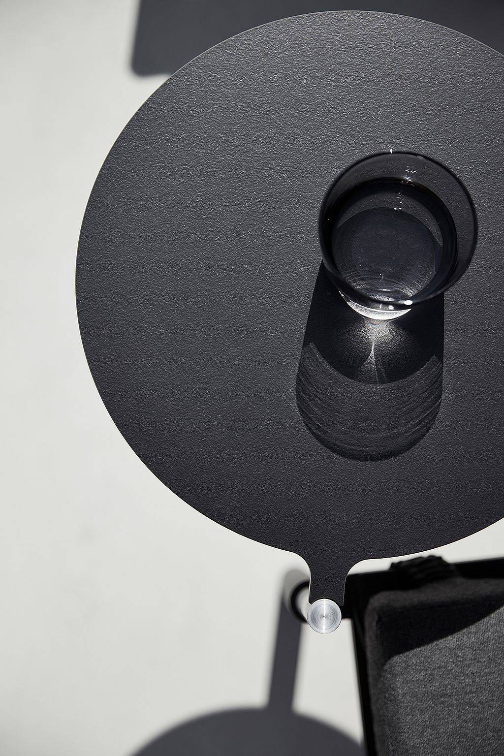Round dark side table attached to a lounge chair, viewed from above with a clear drinking glass casting a sharp shadow on the tabletop.