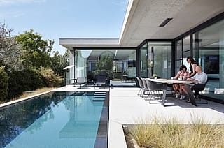 Three people gathered at a dark outdoor dining table beside a narrow pool, outside a modern home with floor-to-ceiling glass and clean concrete lines.