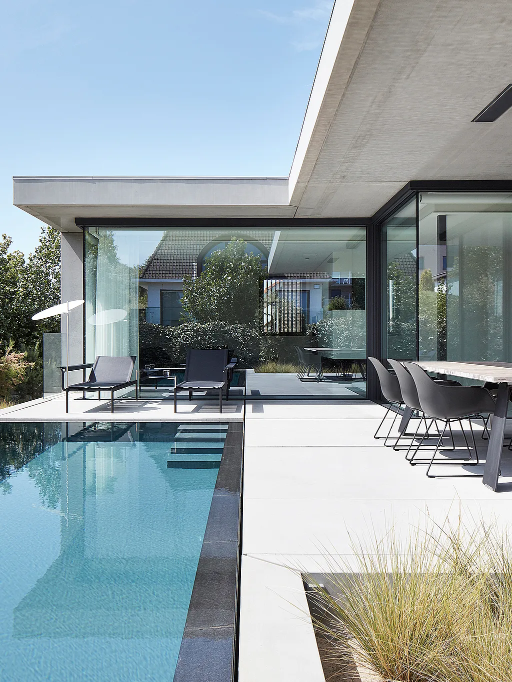 Maison moderne aux murs de verre au bord d’une longue piscine étroite, avec une table à manger extérieure sombre et des chaises sur une terrasse en pierre pâle et deux transats sous un parasol blanc bas à l’extrémité.