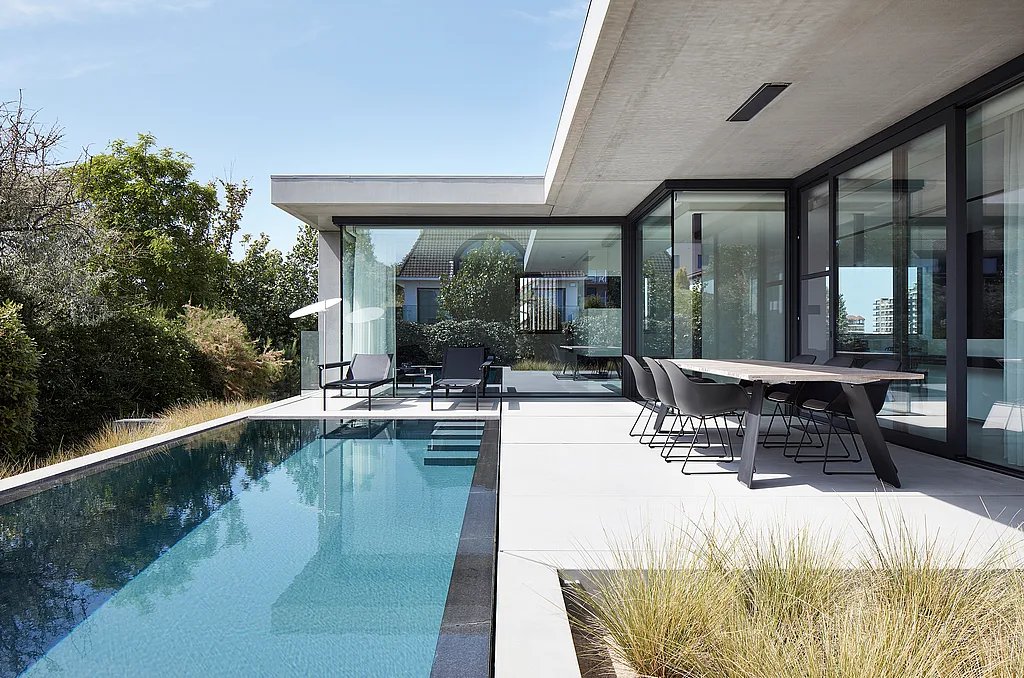 Modernes Haus mit Glaswänden neben einem langen, schmalen Swimmingpool, mit einem dunklen Esstisch und Stühlen im Freien auf einer hellen Steinterrasse und zwei Liegen unter einem niedrigen weißen Sonnenschirm am anderen Ende.