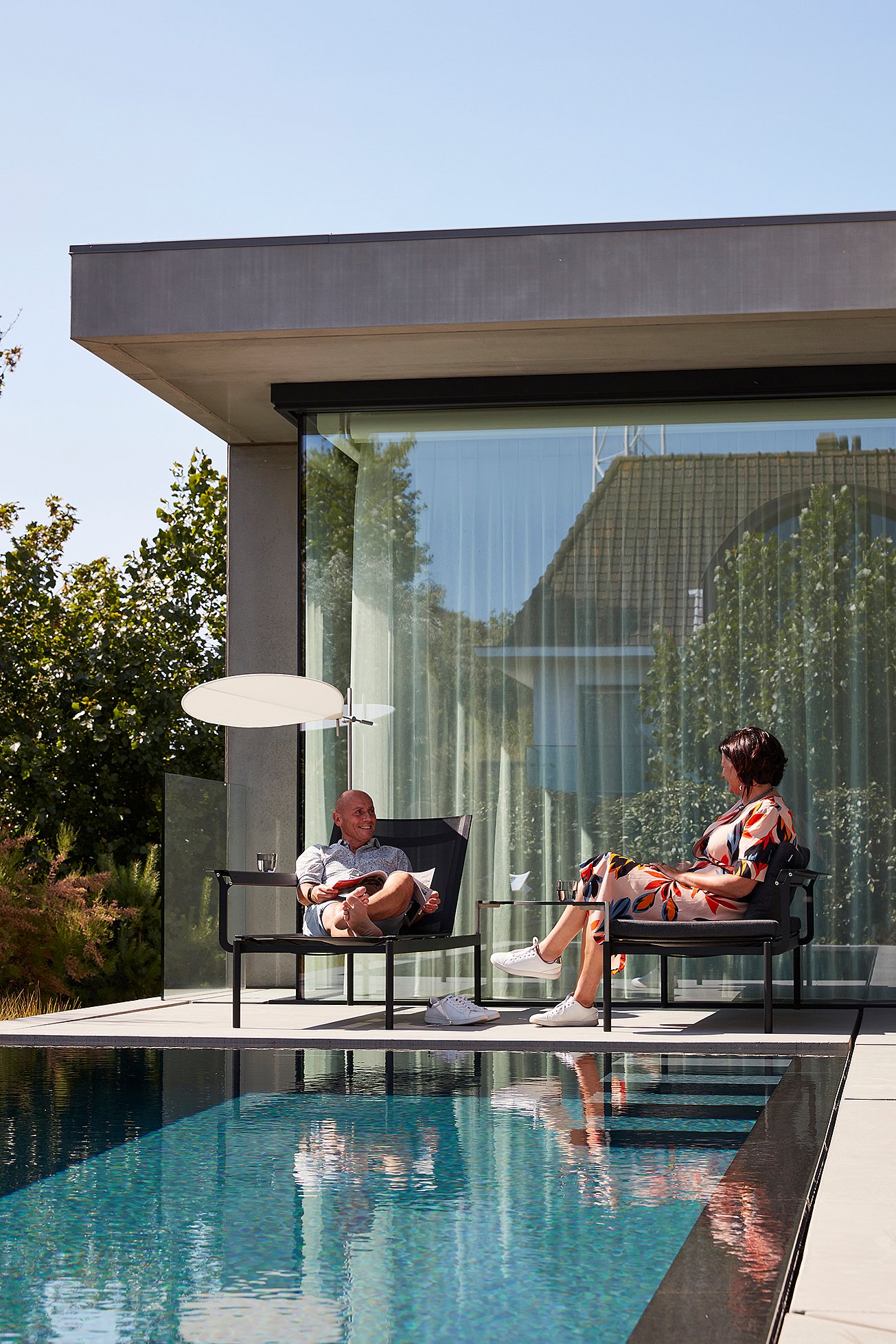 Man and woman relaxing in dark Sol+Luna loungers beside a swimming pool, set against a modern glass façade with a low white parasol between them.