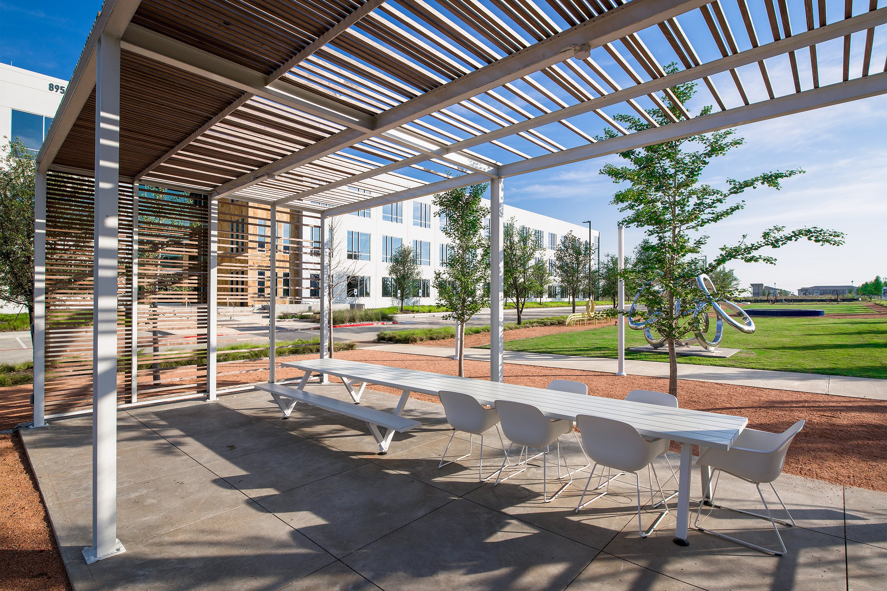 Long white Marina table with white Captain’s Chairs beneath a slatted pergola, facing a modern office façade and a grassy campus with trees and sculpture.