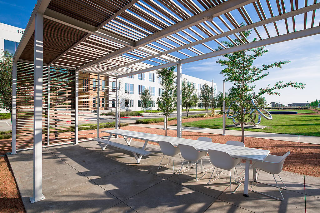 Long white Marina table with white Captain’s Chairs beneath a slatted pergola, facing a modern office façade and a grassy campus with trees and sculpture.
