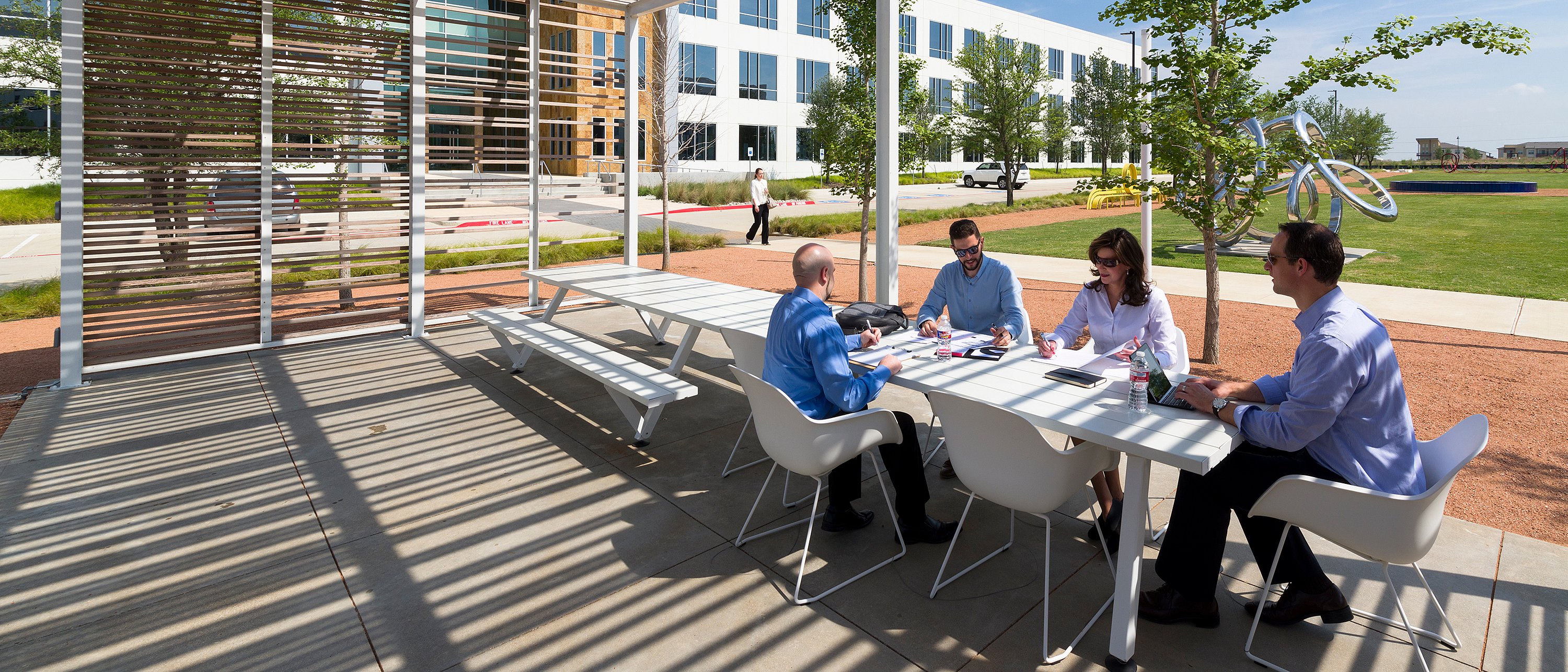 Four colleagues meeting at a white outdoor Marina table under a pergola, with striped shadows across the concrete floor and landscaped office grounds behind them.