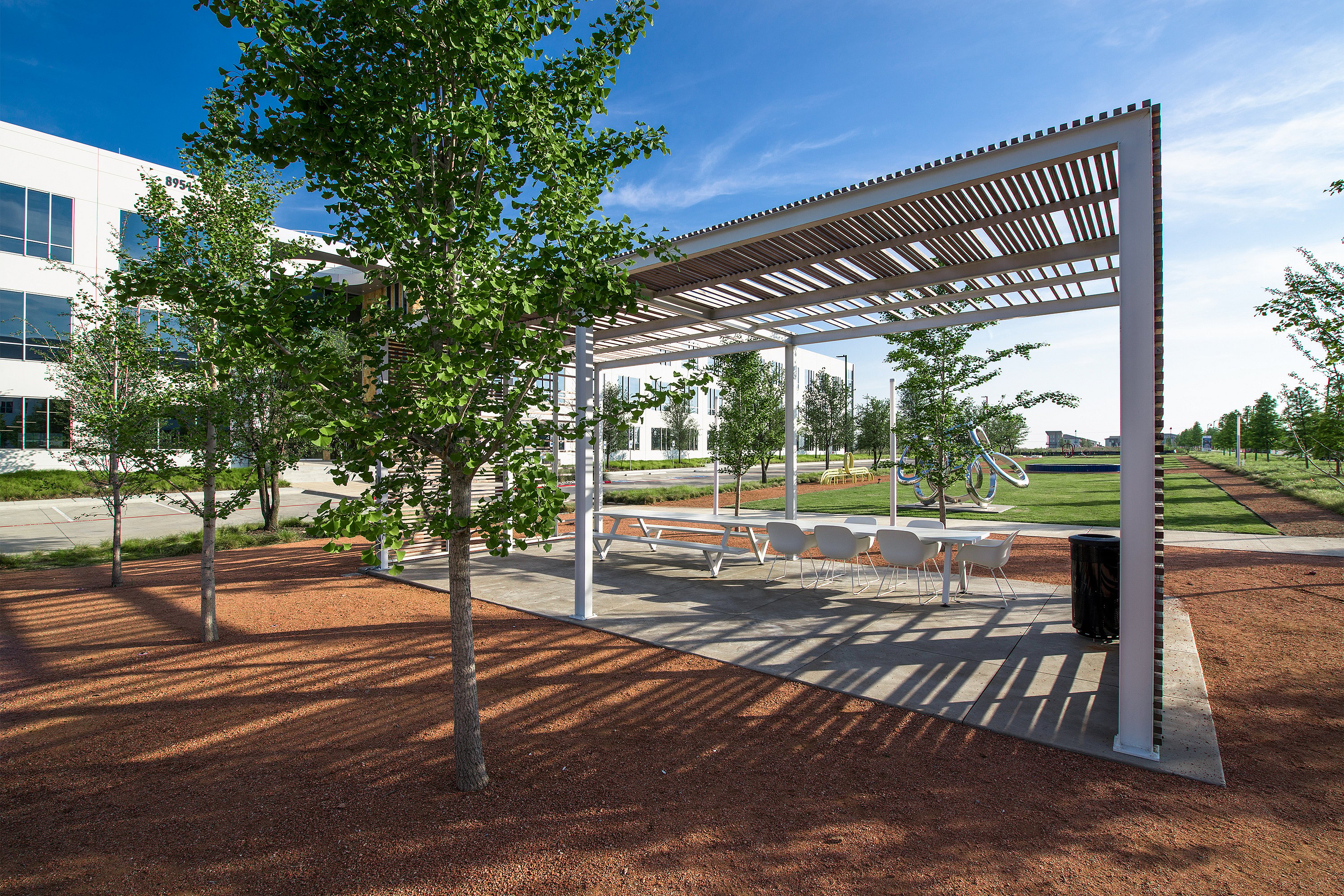 White Marina table with white Captain’s Chairs under a slatted pergola beside a modern office building, surrounded by young trees, red gravel, and a sunny lawn.