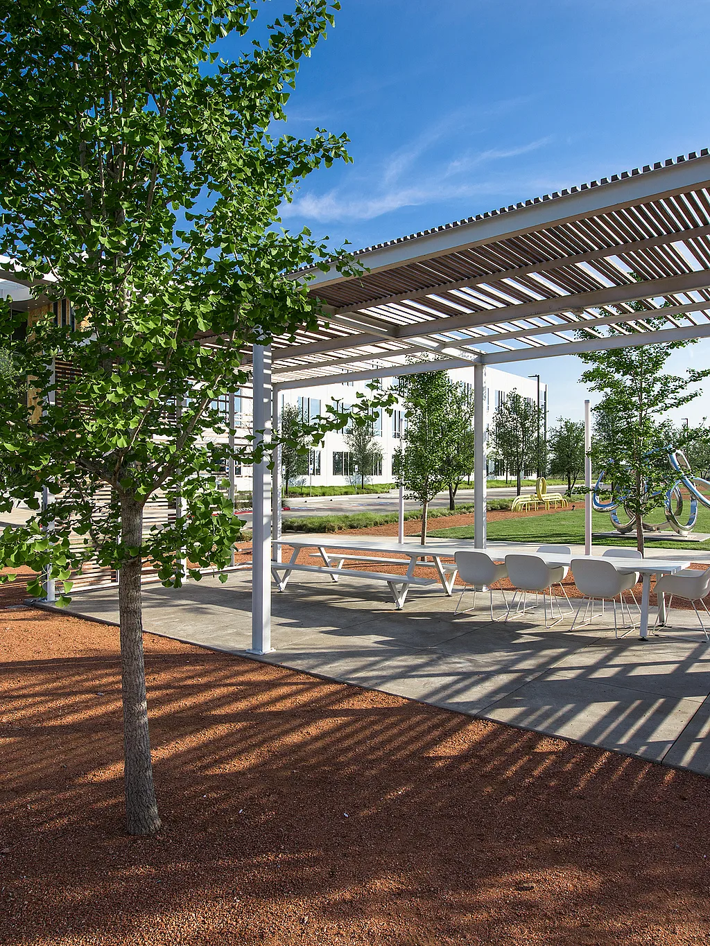 Table de Marina blanche avec des chaises du capitaine blanches sous une pergola à lattes à côté d’un immeuble de bureaux moderne, entouré de jeunes arbres, de gravier rouge et d’une pelouse ensoleillée.