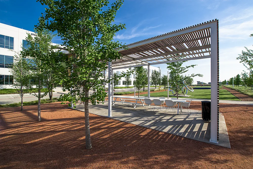 White Marina table with white Captain’s Chairs under a slatted pergola beside a modern office building, surrounded by young trees, red gravel, and a sunny lawn.