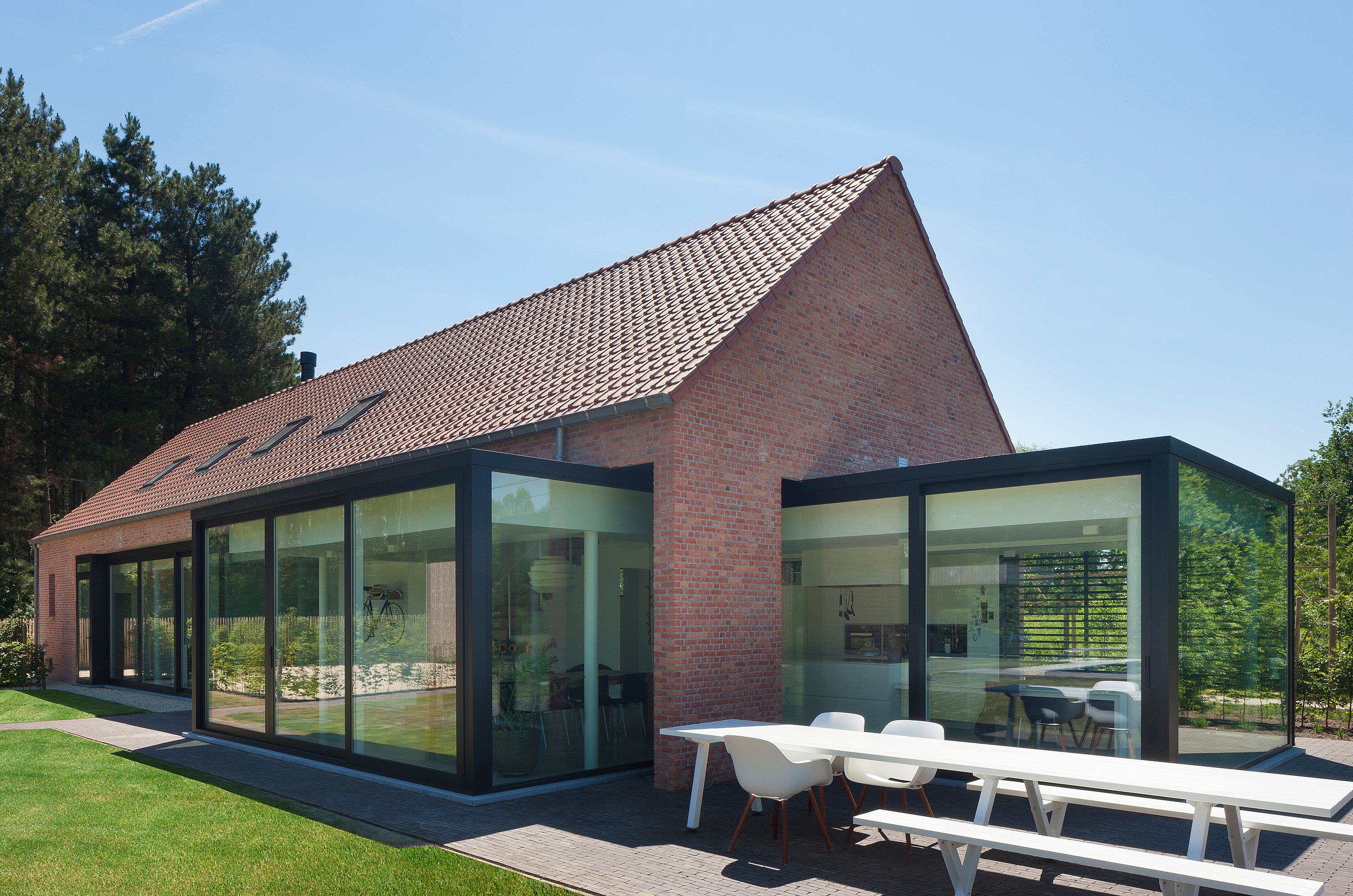 Gerenoveerde bakstenen boerderij met zwarte glasverlengingen, een witte Marina tafel met banken en witte Captain's stoelen op het terras en een zonovergoten gazon op de voorgrond.
