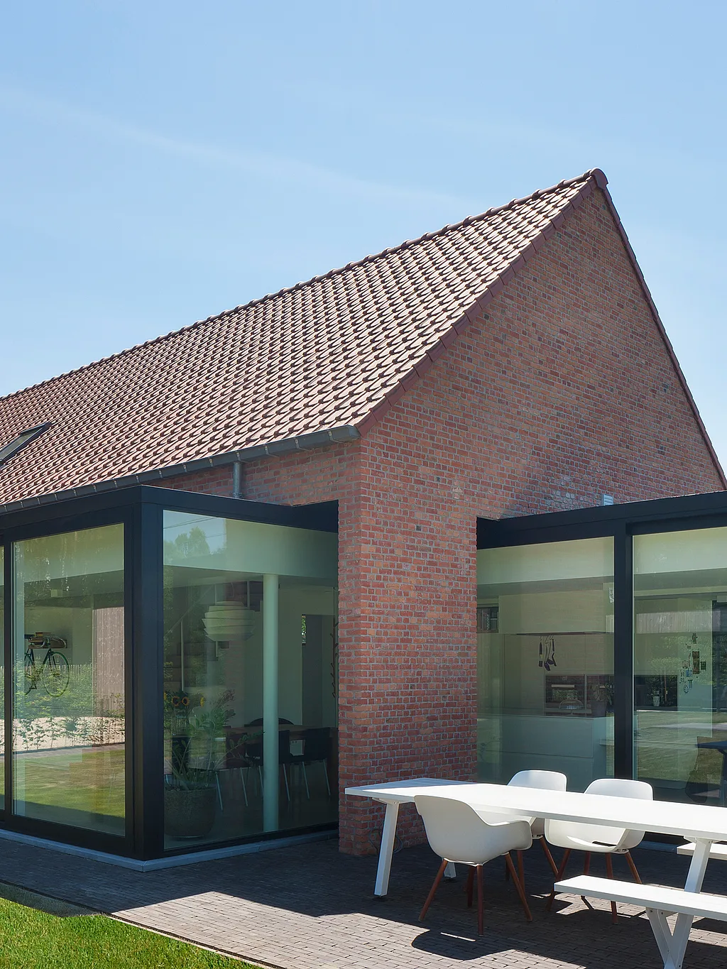 Gerenoveerde bakstenen boerderij met zwarte glasverlengingen, een witte Marina tafel met banken en witte Captain's stoelen op het terras en een zonovergoten gazon op de voorgrond.