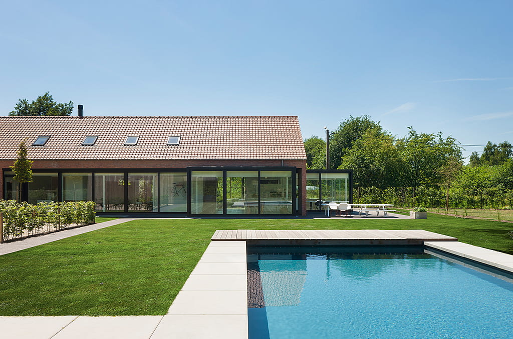 Wide garden view of the renovated farmhouse with a long glass façade, a rectangular swimming pool, manicured lawn, and the white Marina dining set on the terrace to the right.