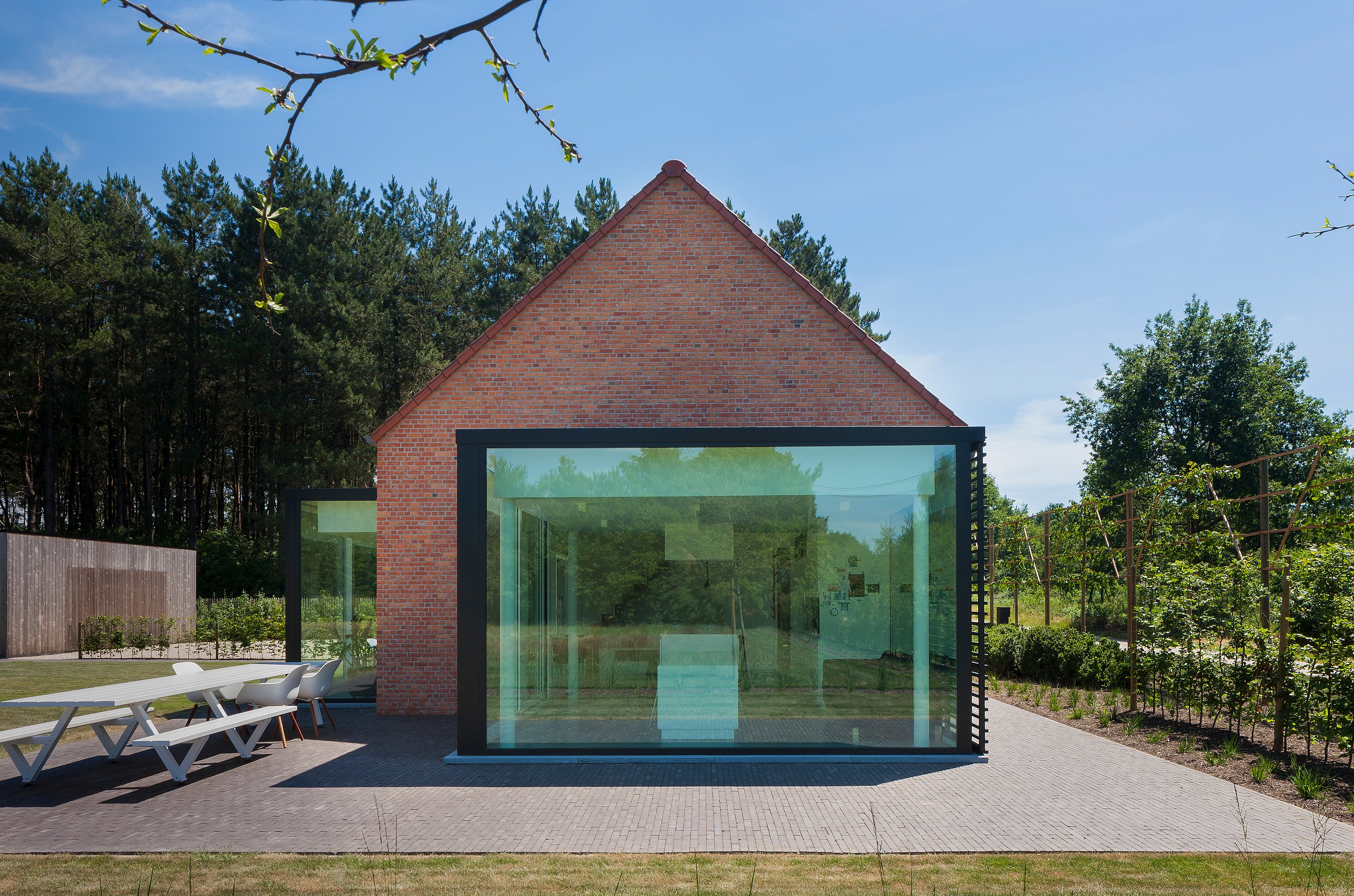 Front view of the brick farmhouse with a large black-framed glass extension, surrounded by trees and garden planting, with the white Marina table and chairs visible on the paved terrace.