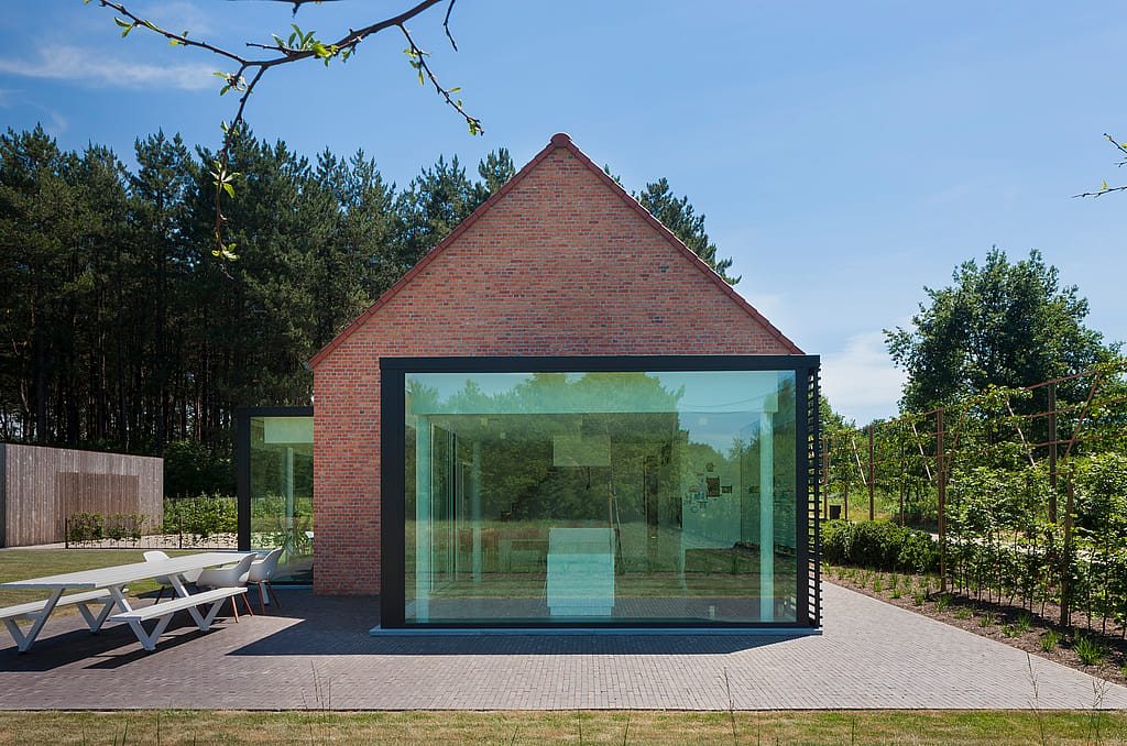 Front view of the brick farmhouse with a large black-framed glass extension, surrounded by trees and garden planting, with the white Marina table and chairs visible on the paved terrace.