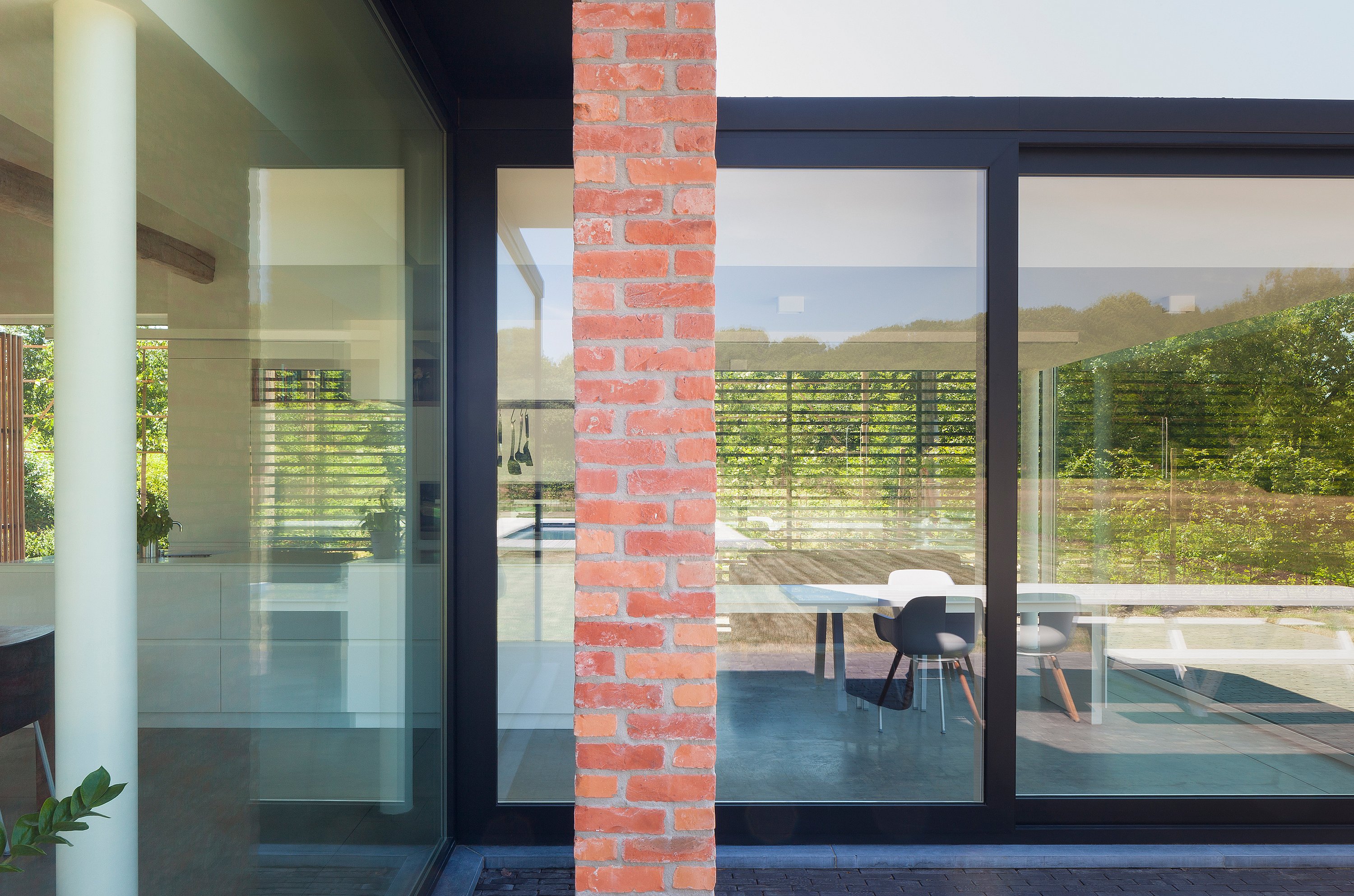 Close view through black-framed glass windows divided by a brick column, showing the interior and the white Marina table with mixed white and dark chairs reflected through the façade.