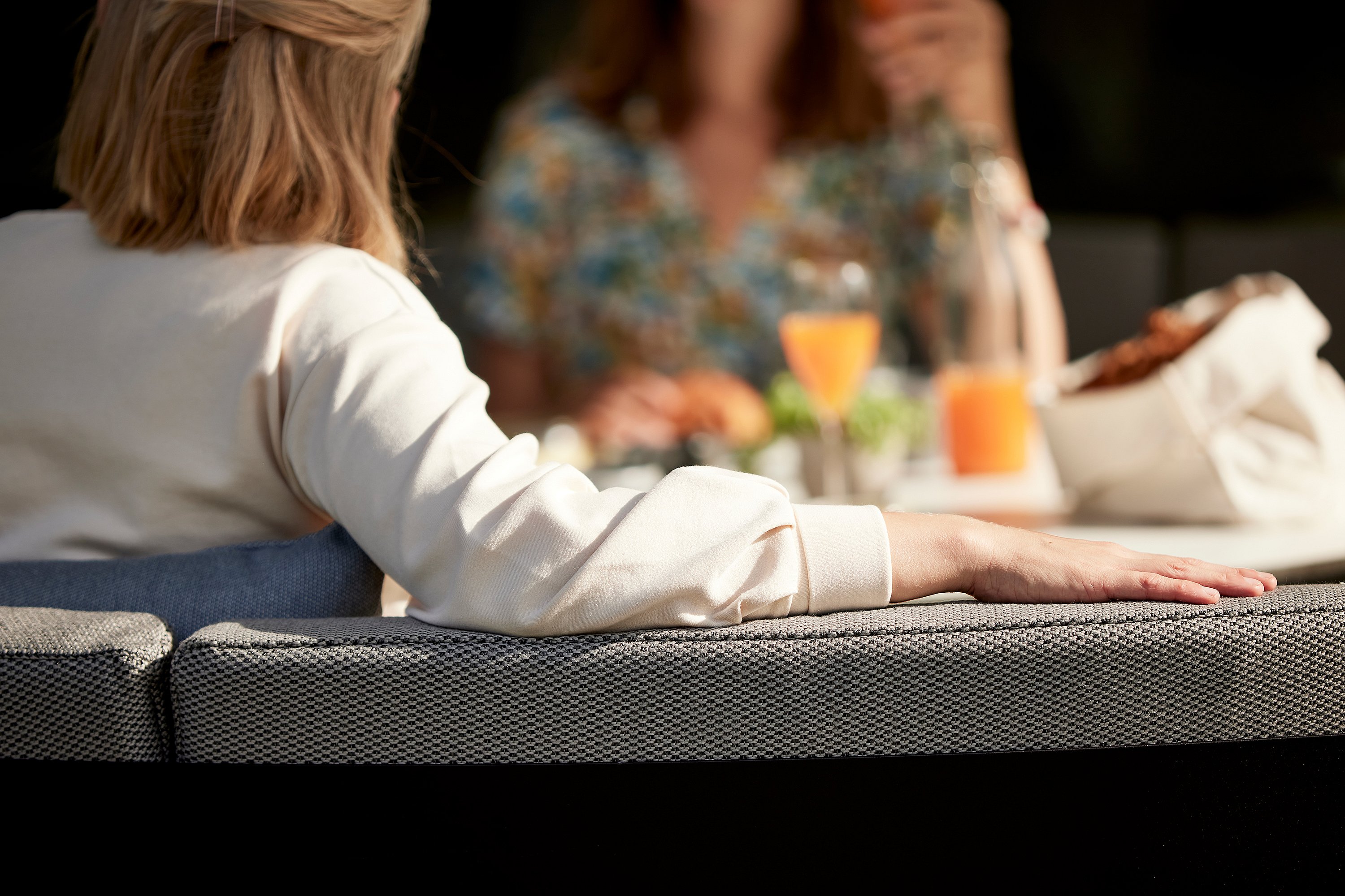 Graues gewölbtes Kosmos-Außensofa mit einem weißen runden Tisch auf einer überdachten Terrasse, durch offene Schiebetüren mit dem Garten dahinter betrachtet.