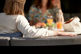 Graues gewölbtes Kosmos-Außensofa mit einem weißen runden Tisch auf einer überdachten Terrasse, durch offene Schiebetüren mit dem Garten dahinter betrachtet.
