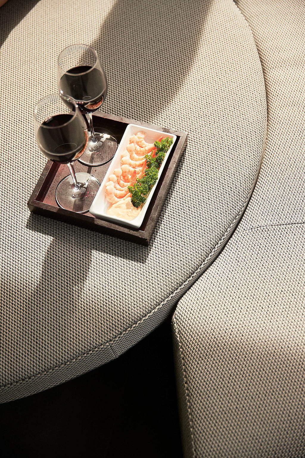 Nahaufnahme von zwei Gläsern Rotwein und einer Garnelen-Vorspeise über dem Kopf auf einem Tablett, das auf dem hellgrauen Kissen des Kosmos-Sofas steht.