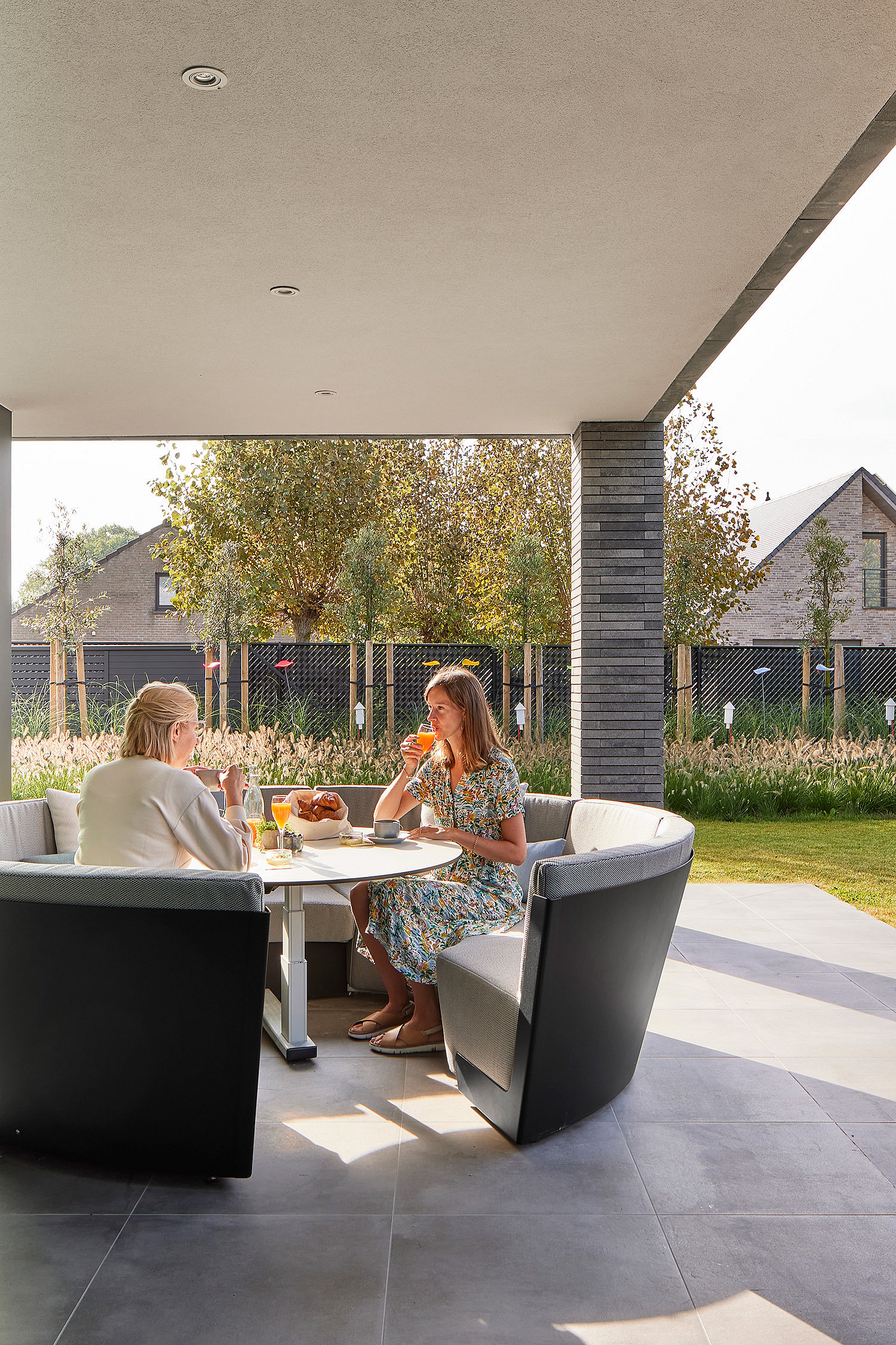 Twee vrouwen ontbijten aan een witte ronde Kosmos-tafel in een gebogen grijze buitenbank op een overdekt terras, met een gazon en naburige huizen op de achtergrond.