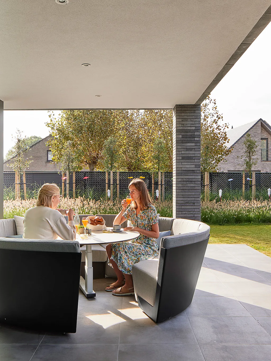 Two women having breakfast at a white round Kosmos table inside a curved gray outdoor sofa on a covered terrace, with a lawn and neighboring houses in the background.