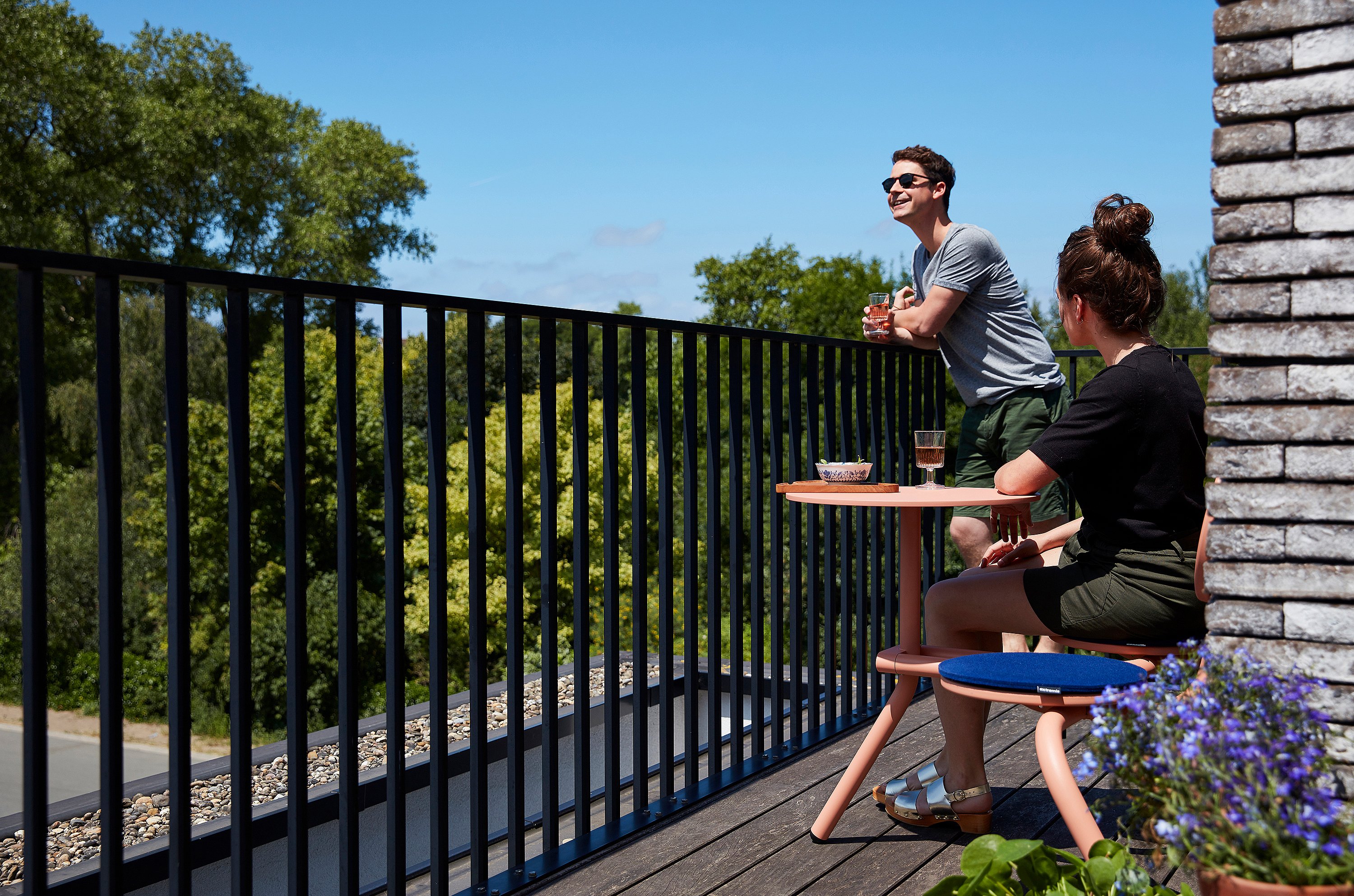 Zwei Personen genießen Getränke auf einem schmalen Balkon, wobei eine Person an einem korallenfarbenen Bistroo-Tisch sitzt und die andere sich auf das schwarze Geländer lehnt und unter einem strahlend blauen Himmel auf die grünen Baumwipfel blickt.