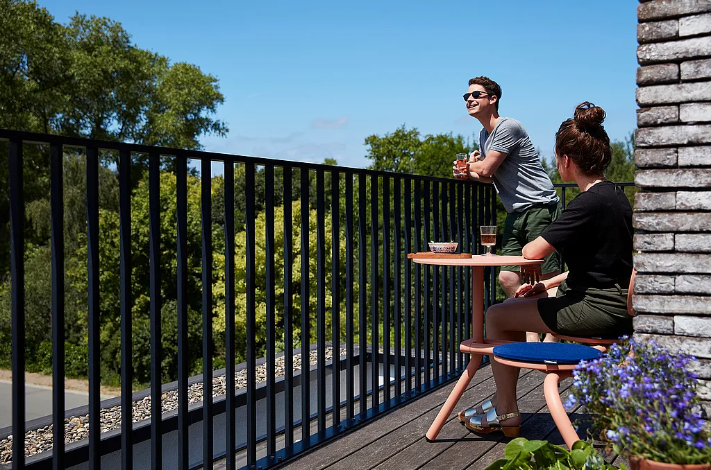 Two people enjoying drinks on a narrow balcony, with one person seated at a coral Bistroo table and the other leaning on the black railing, overlooking leafy treetops under a bright blue sky.