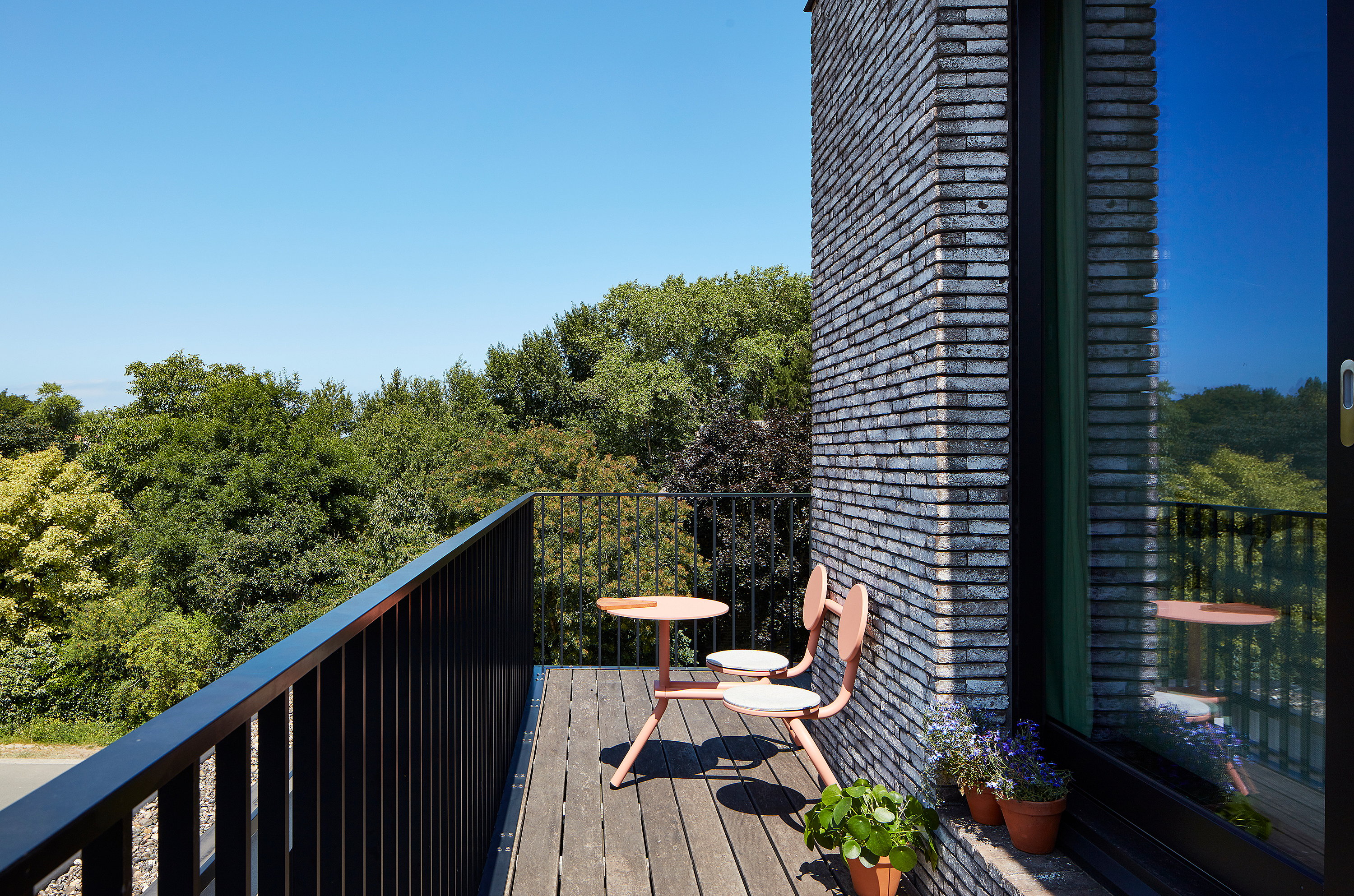 Coral Bistroo balkon met twee stoelen met ronde rug en een kleine ronde tafel op een smal houten balkon naast een bakstenen muur en een reflecterend raam, met bomen voorbij de reling.