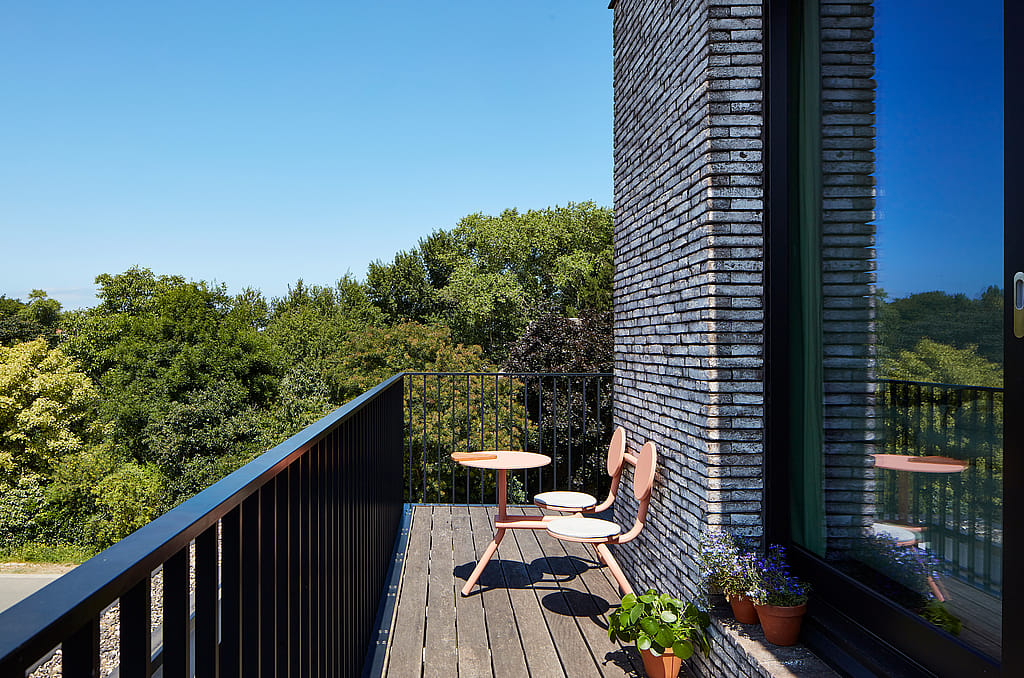 Coral Bistroo balkon met twee stoelen met ronde rug en een kleine ronde tafel op een smal houten balkon naast een bakstenen muur en een reflecterend raam, met bomen voorbij de reling.