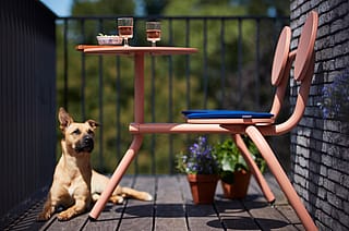 Coral Bistroo balkon tafel en stoel met blauwe zitkussens op een houten dek naast potbloemen, met een hond in de zon bij de reling.