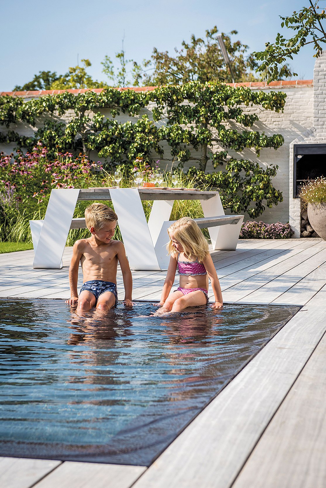 Two children sitting at the edge of an infinity pool on a wooden terrace, with a white Hopper picnic table and lush garden behind them.