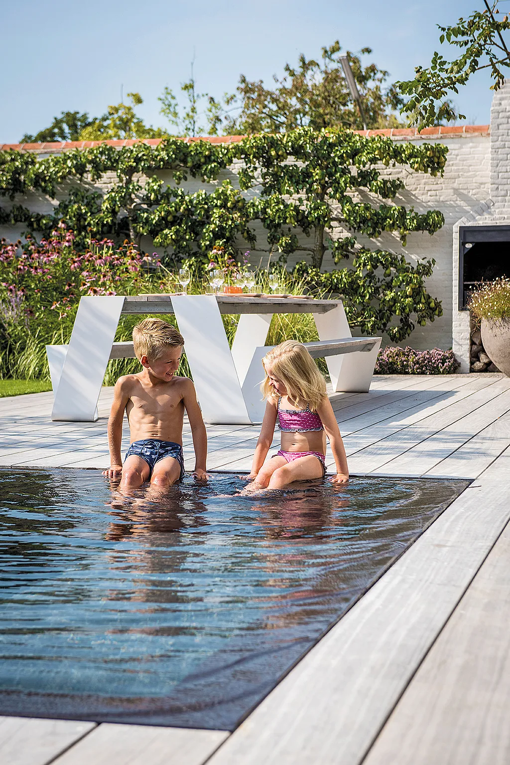 Two children sitting at the edge of an infinity pool on a wooden terrace, with a white Hopper picnic table and lush garden behind them.