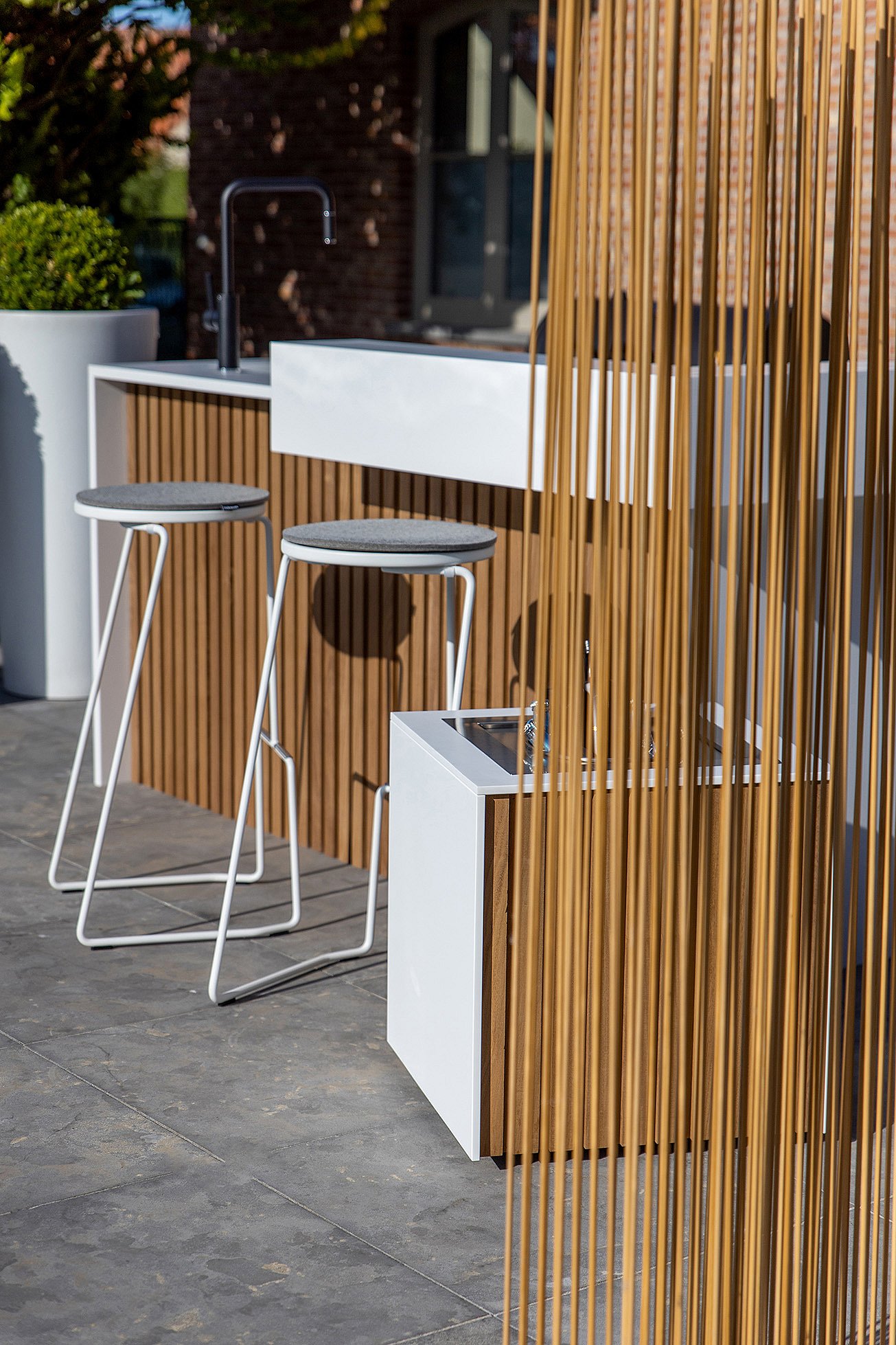 Side view of the outdoor kitchen and white barstools beside a wood-toned Sticks room divider and matching ice bucket on a stone terrace.