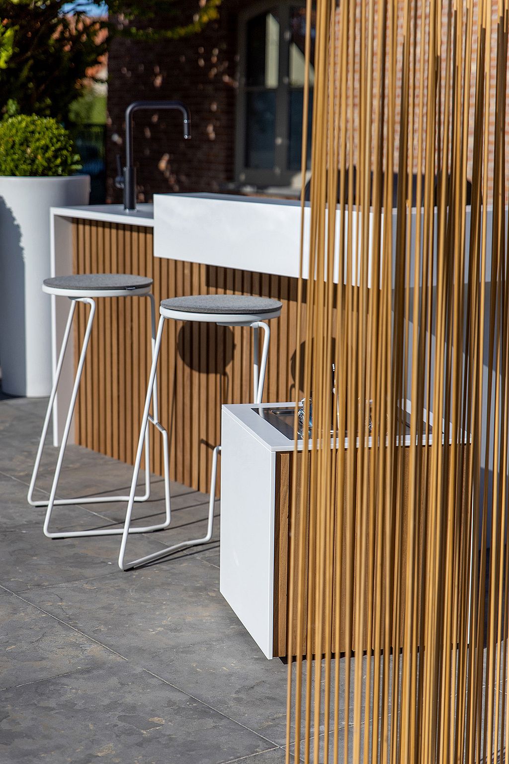 Side view of the outdoor kitchen and white barstools beside a wood-toned Sticks room divider and matching ice bucket on a stone terrace.