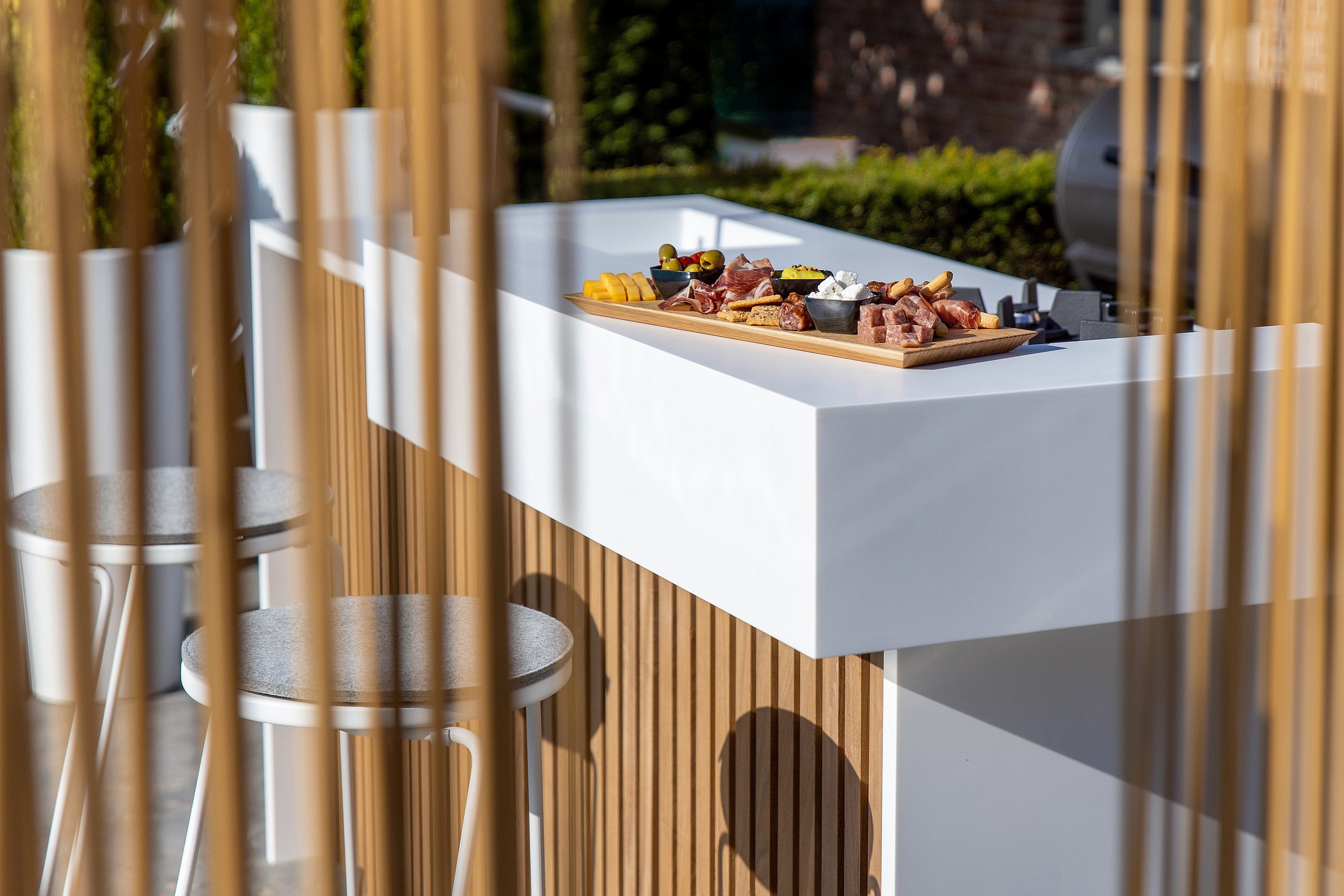 Outdoor kitchen counter seen through vertical copper-toned Sticks, with a serving board of appetizers on the white countertop and tiki barstools in front.