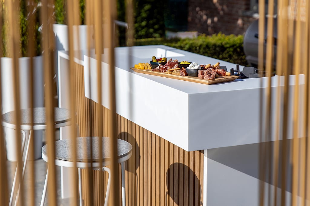 Outdoor kitchen counter seen through vertical copper-toned Sticks, with a serving board of appetizers on the white countertop and tiki barstools in front.