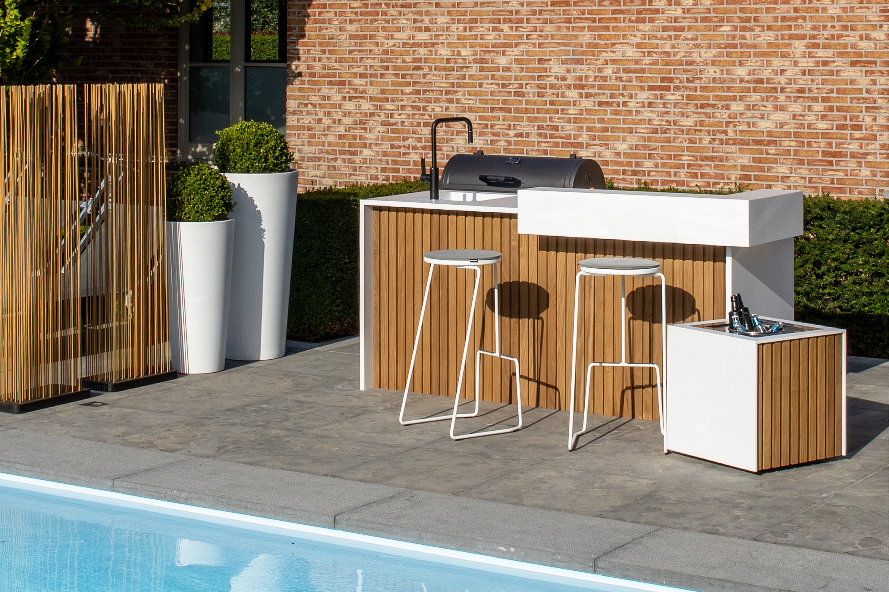 Witte buitenkeuken met houten latten naast een zwembad, met twee witte tiki barkrukken, hoge plantenbakken en een koperkleurige Sticks-scherm voor een bakstenen huis.