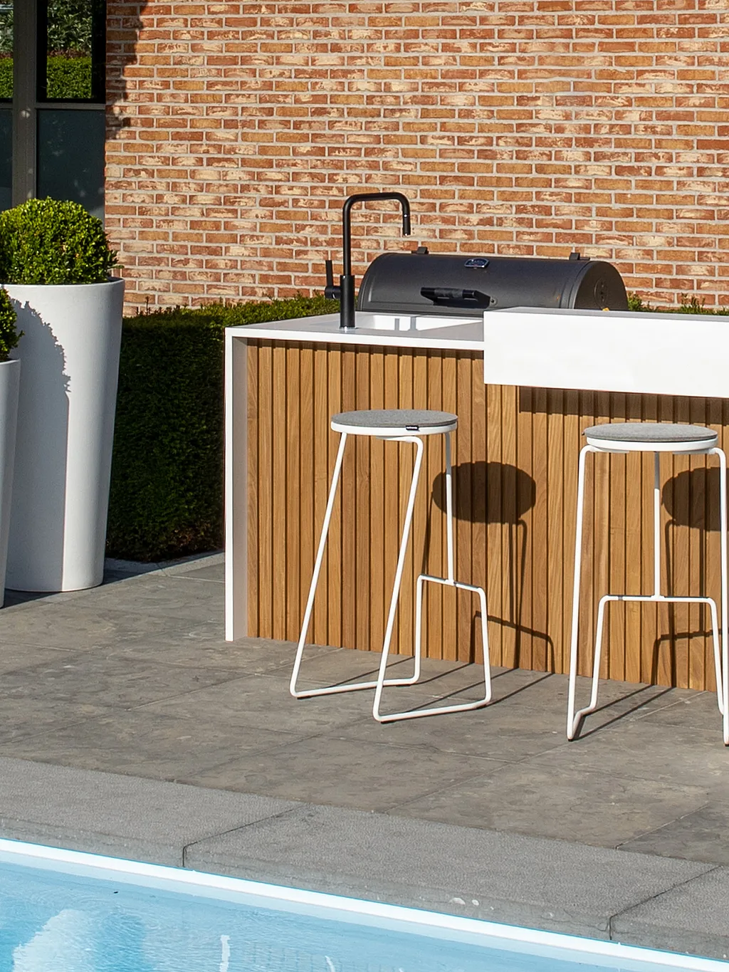 Witte buitenkeuken met houten latten naast een zwembad, met twee witte tiki barkrukken, hoge plantenbakken en een koperkleurige Sticks-scherm voor een bakstenen huis.